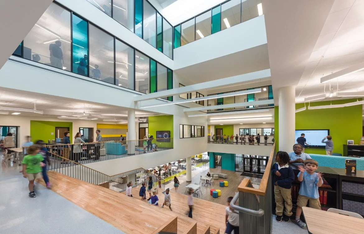 Interior view of a multi-story modern school or educational facility with students and staff, featuring open spaces, glass walls, and colorful walls in green and yellow, with children on the staircase and adults in conversation.