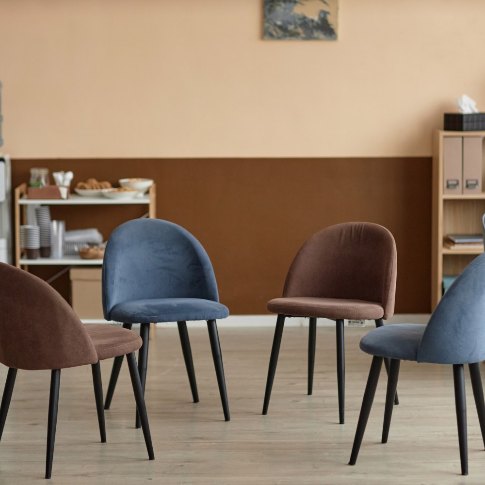 A room with five upholstered chairs in blue and brown, arranged in a semi-circle on a wood floor, with a shelf of dishes and a wall art in the background.