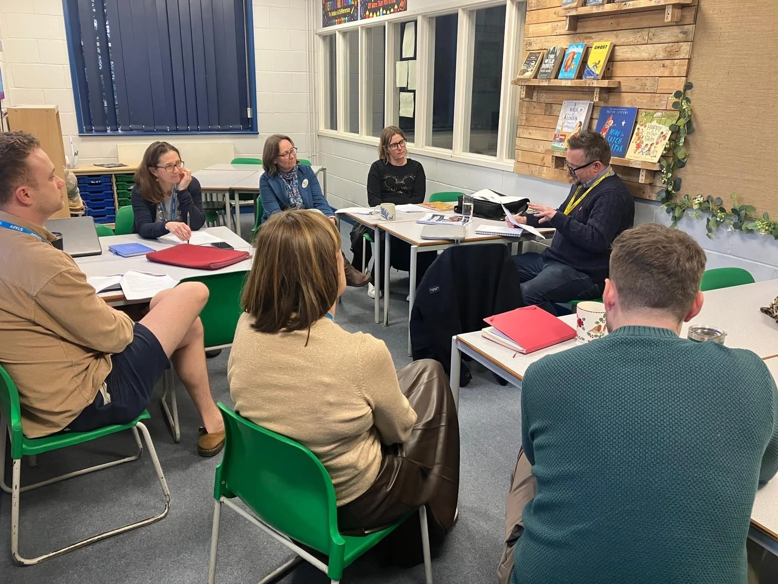 A group of adults attending a meeting or workshop in a classroom-like setting, seated around tables. There is a man at the front reading or presenting, with books and notebooks on the tables. The room has a window, some books on a wall shelf, and decorative greenery.