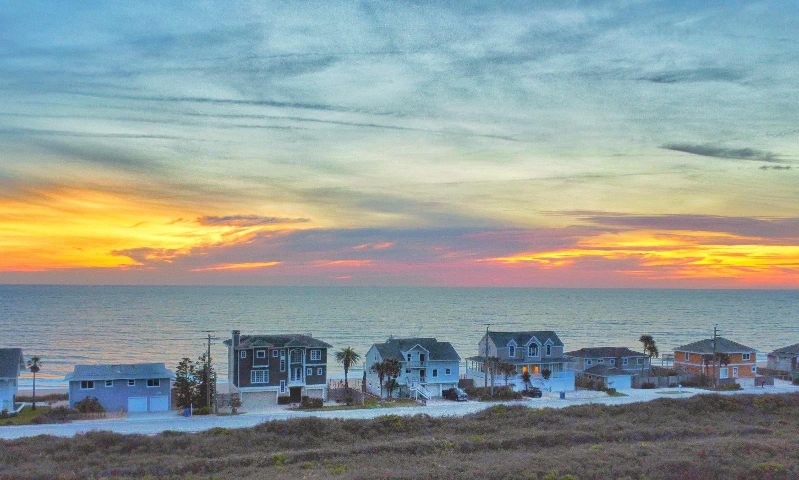 A row of beach houses along the shoreline at sunset with the ocean in the background, colorful sky with clouds.