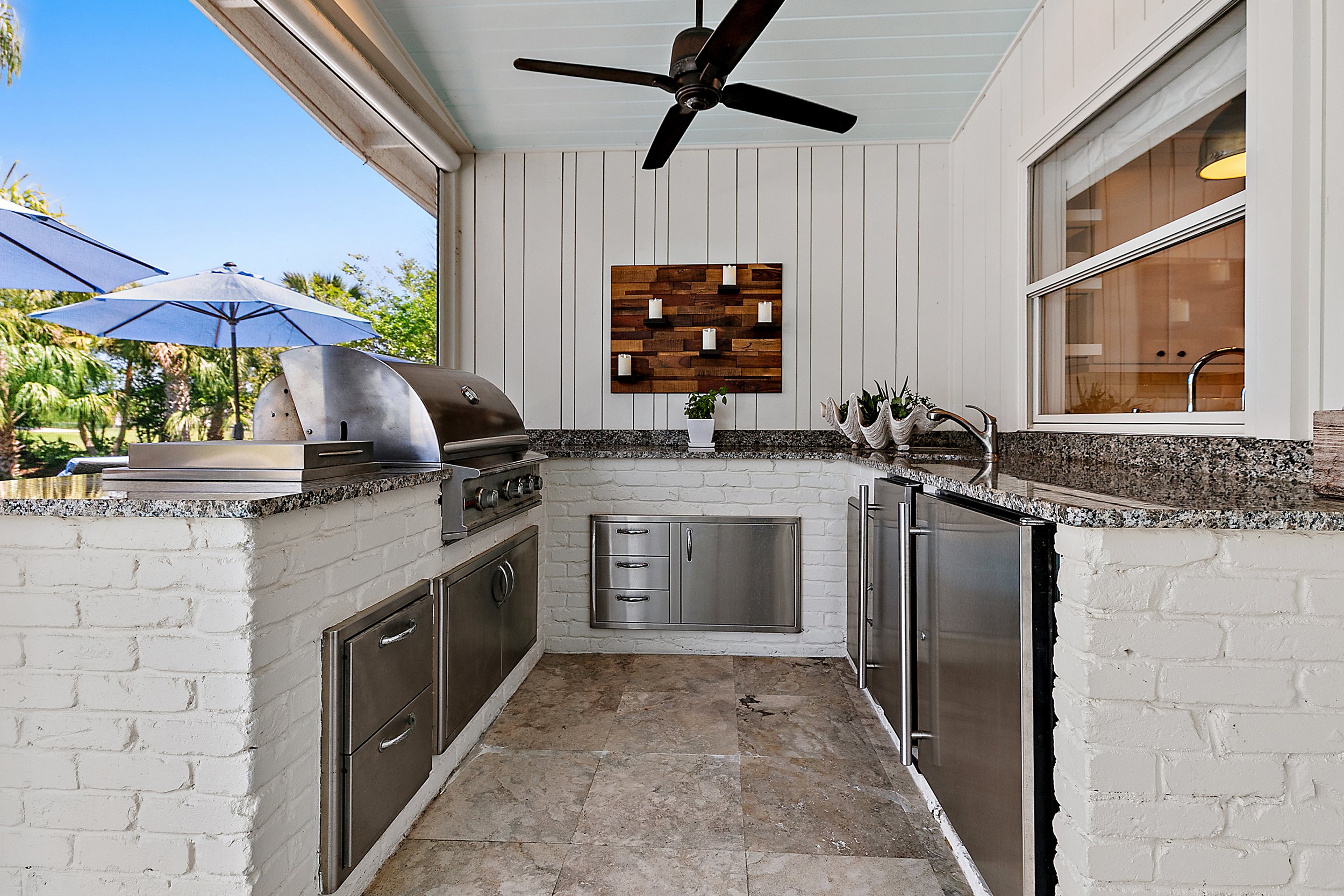 Outdoor kitchen with stainless steel appliances, granite countertops, white brick walls, a ceiling fan, and a window overlooking a garden with umbrellas.