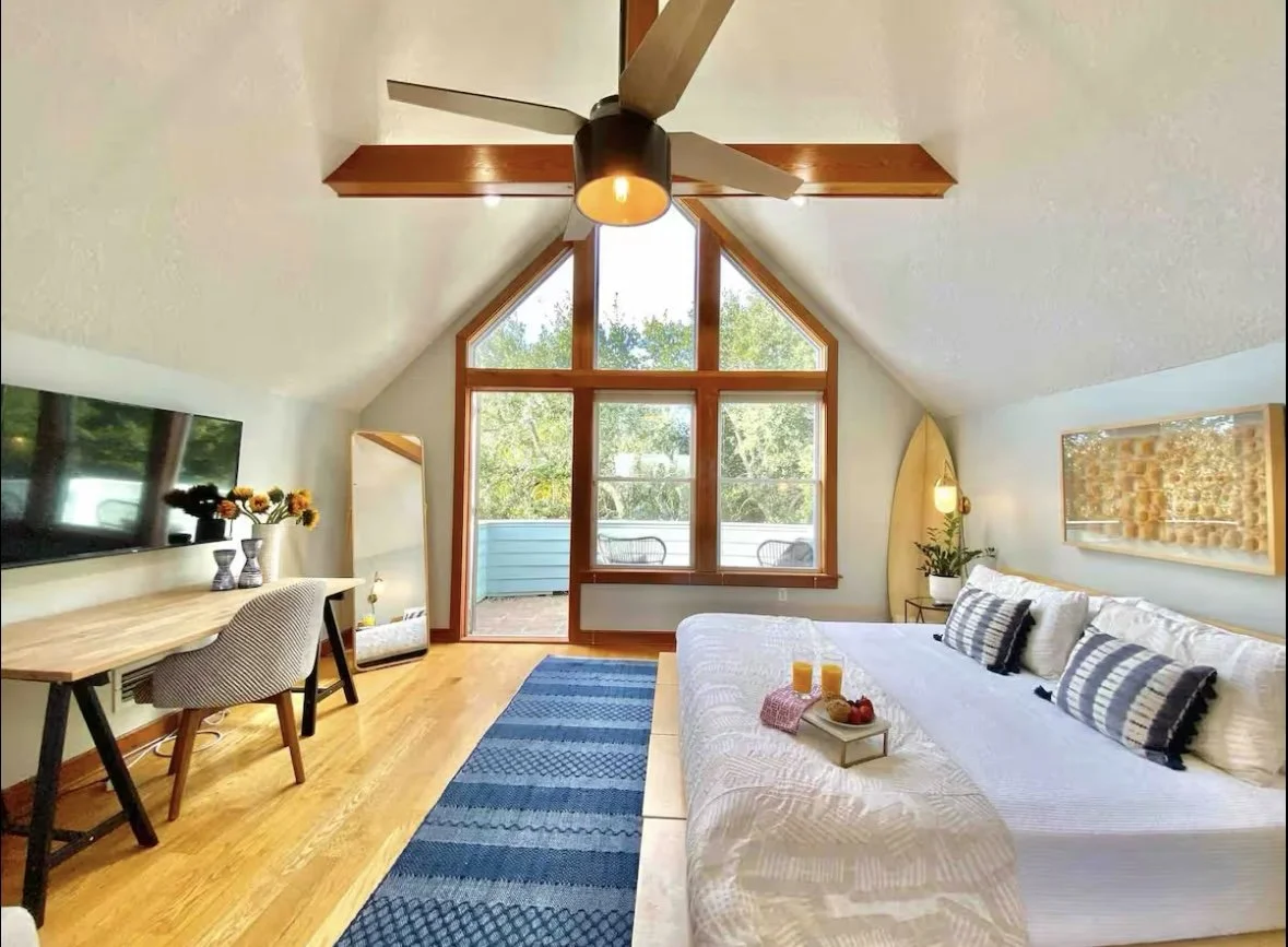 Bright bedroom with large window and pitched ceiling, featuring a bed with striped pillows, a wooden desk with a chair, a wall mirror, a hanging surfboard, and a sliding glass door leading to a balcony with outdoor chairs.