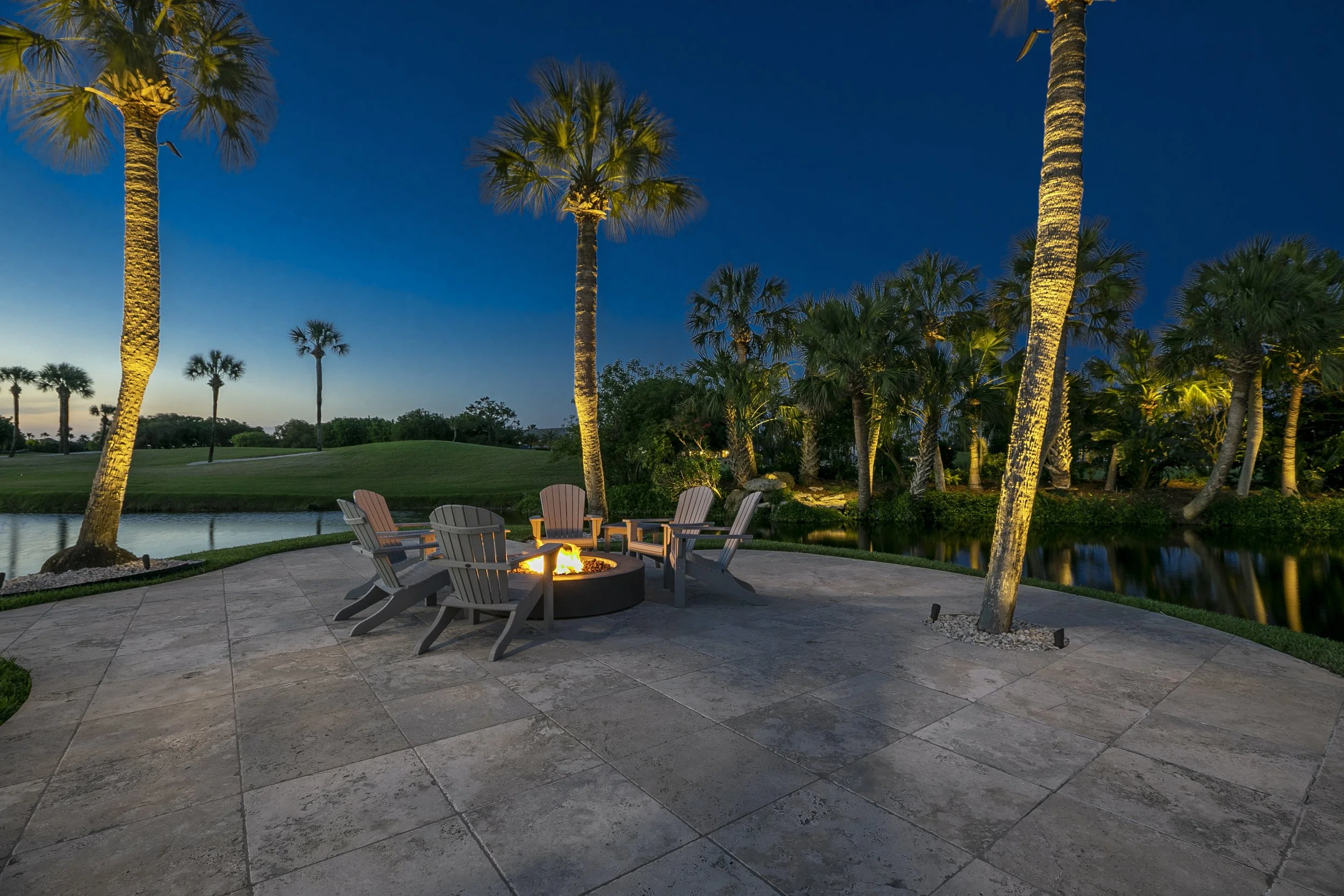 A backyard at dusk with a fire pit surrounded by Adirondack chairs, tall palm trees, a pond, and a grassy landscape.