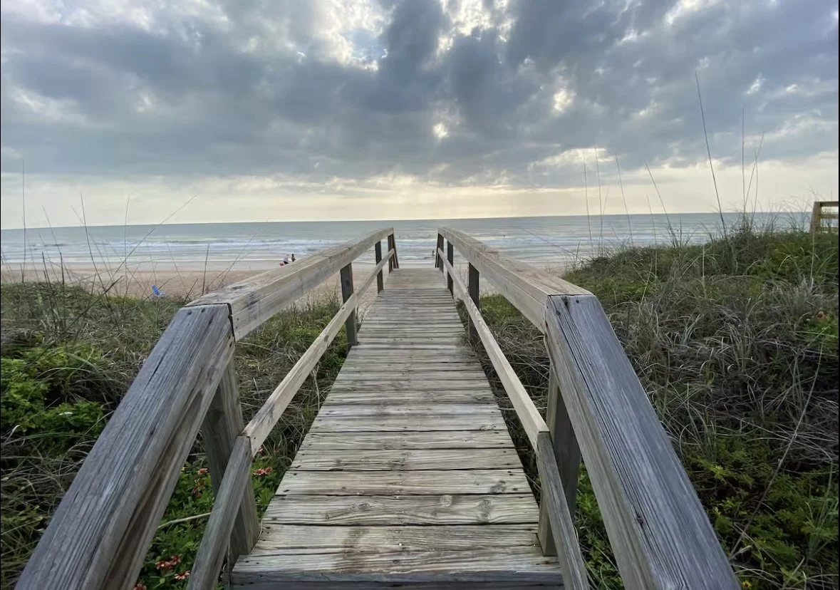 Wooden boardwalk leading to a sandy beach with ocean waves and cloudy sky.