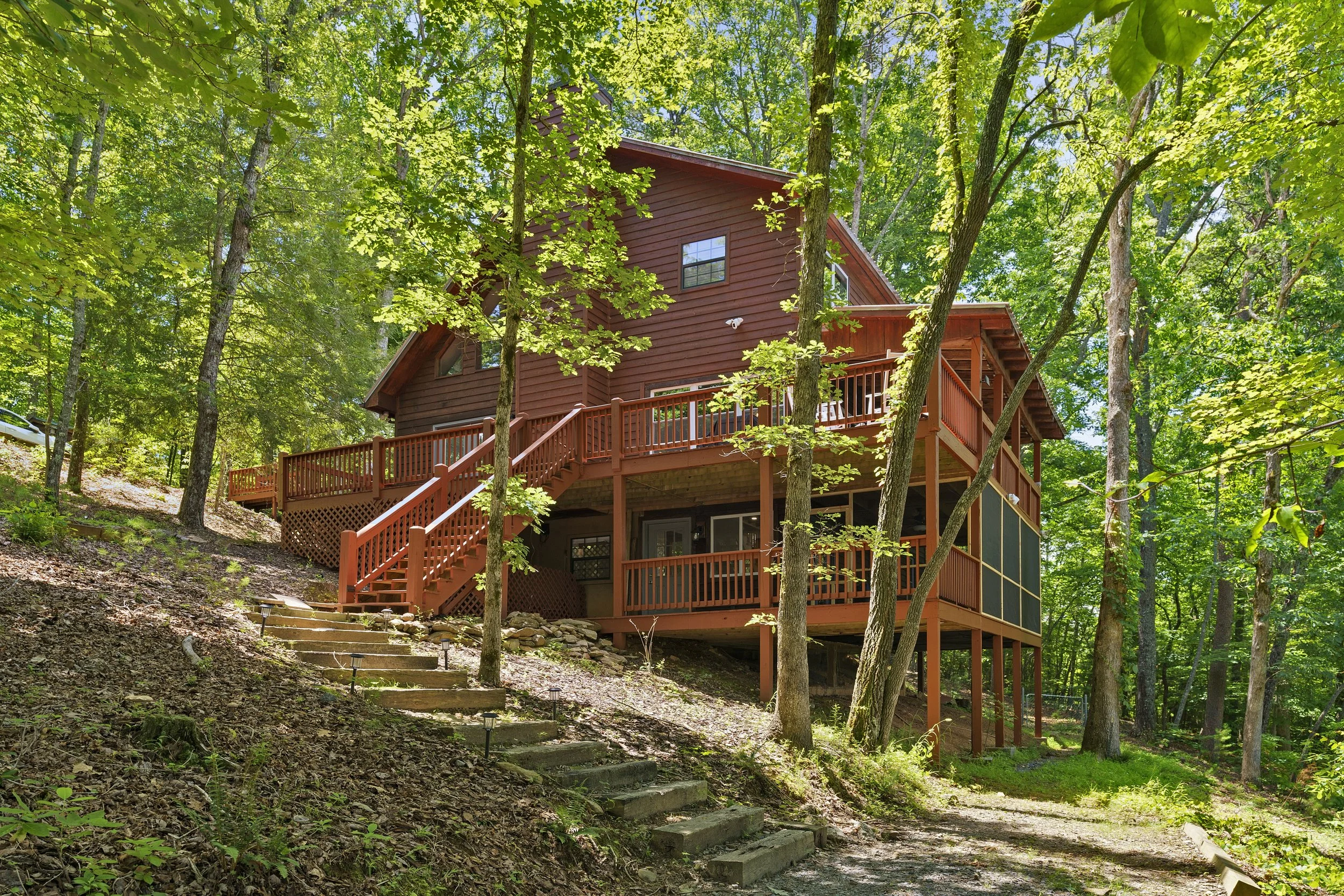 A multi-story wooden house with a red exterior is situated among tall green trees. It has multiple decks with wooden railings and a staircase leading up to the entrance. Sunlight filters through the leaves, illuminating the natural wooded setting.