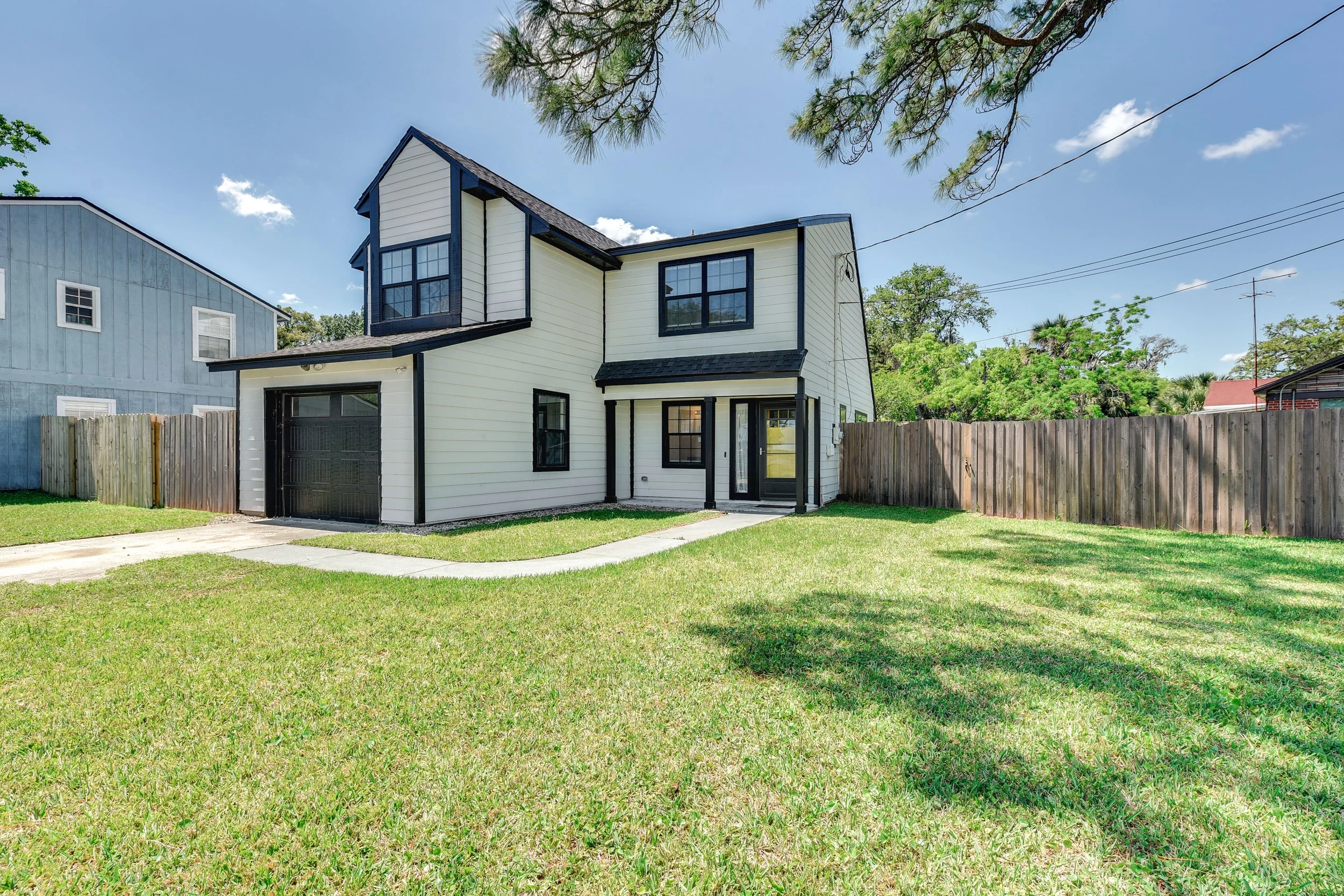 Front view of a modern two-story house with white siding, black trim, a small front porch, and a garage, surrounded by a grassy yard and a wooden fence.