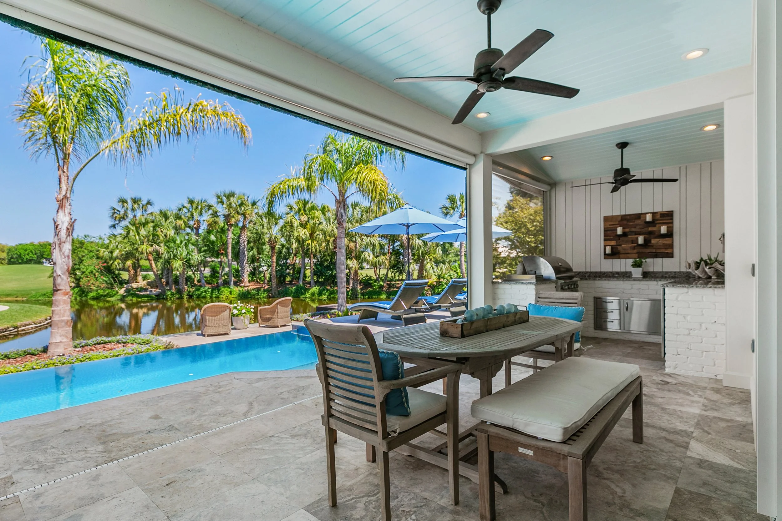 Indoor-outdoor living space with a covered patio, ceiling fans, a dining table with chairs and a bench, an outdoor kitchen with a grill, and a view of a backyard with a pool, palm trees, and lounge chairs with umbrellas.