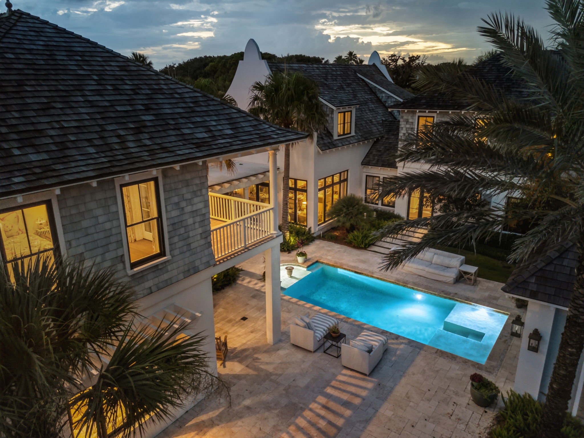 Aerial view of a modern house with lit windows, a swimming pool, and outdoor seating at dusk, surrounded by palm trees and lush greenery.