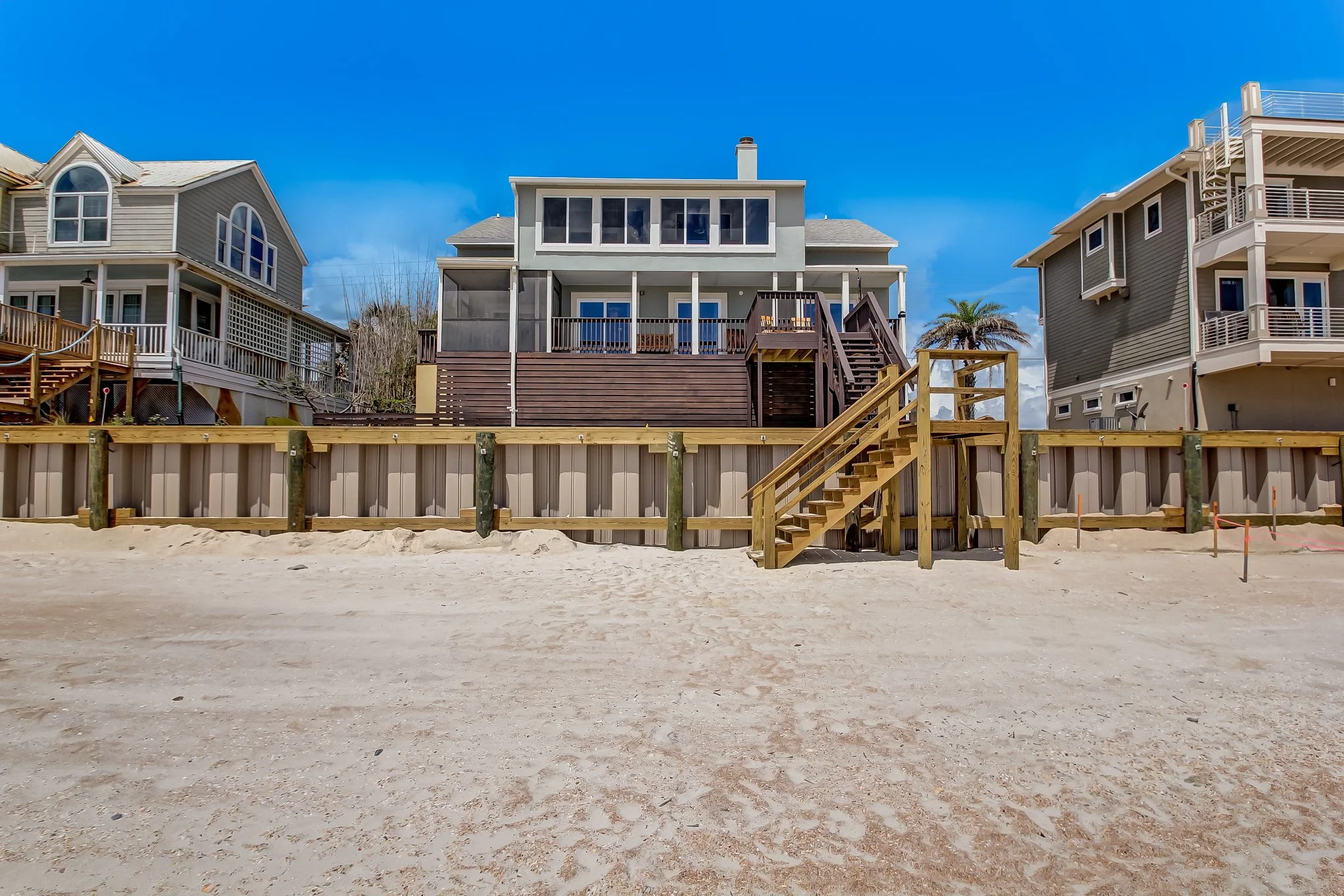 Beachfront houses with decks, stairs, and sand in the foreground under a clear blue sky.