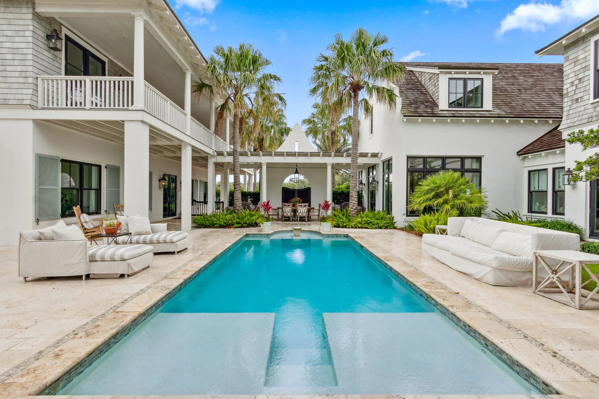 Luxury backyard with a swimming pool, outdoor seating, palm trees, and a white house with black windows and a balcony