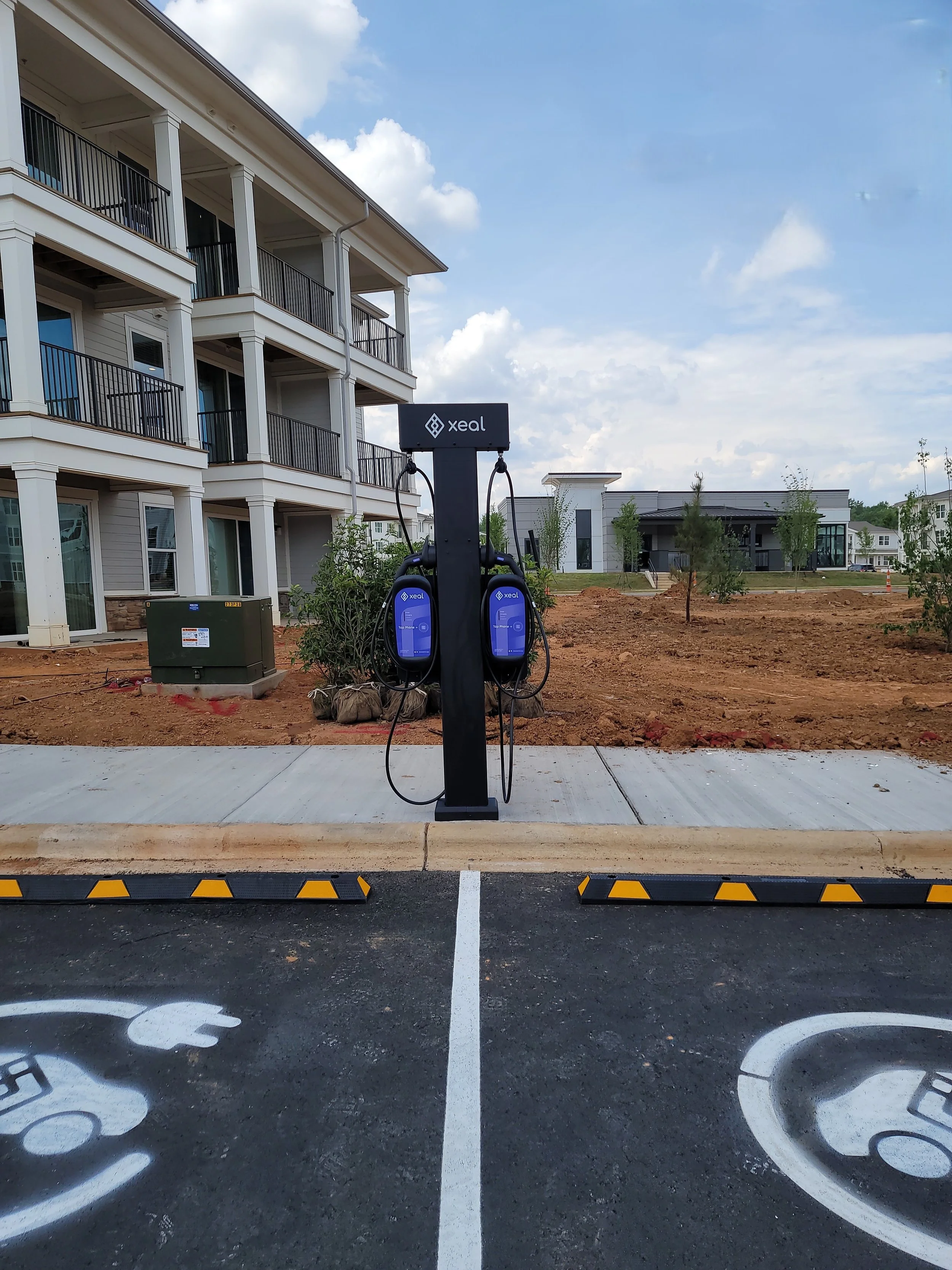 Electric vehicle charging station labeled 'xeal' in a parking lot, with apartment buildings and a partly cloudy sky in the background.