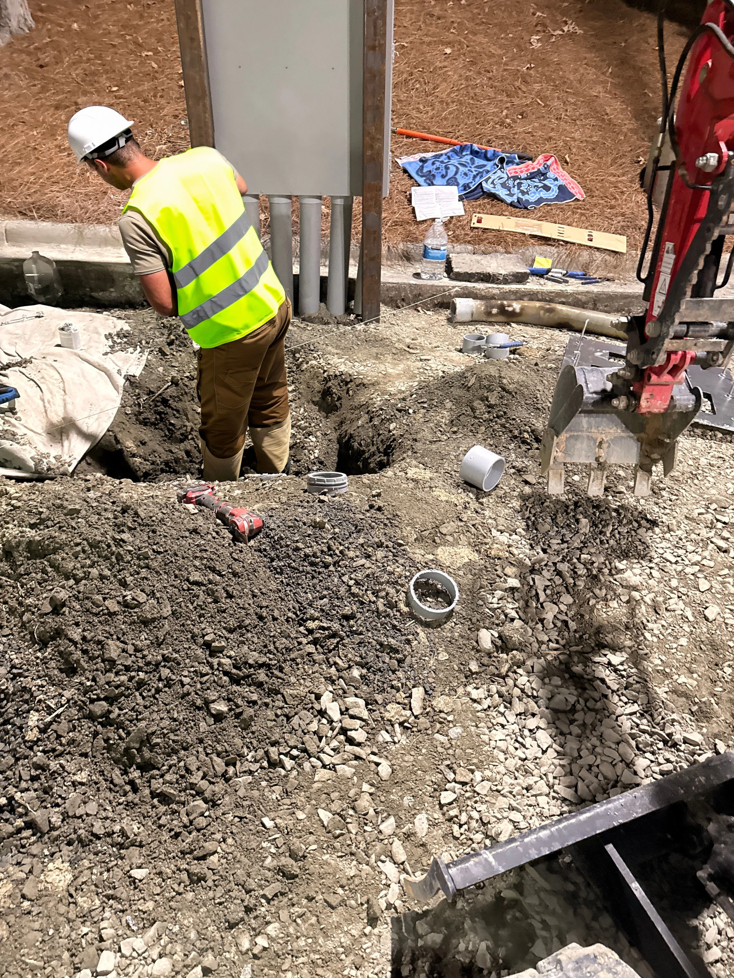 Construction worker in a high-visibility vest and hard hat working at an excavation site with pipes, tools, and construction equipment.