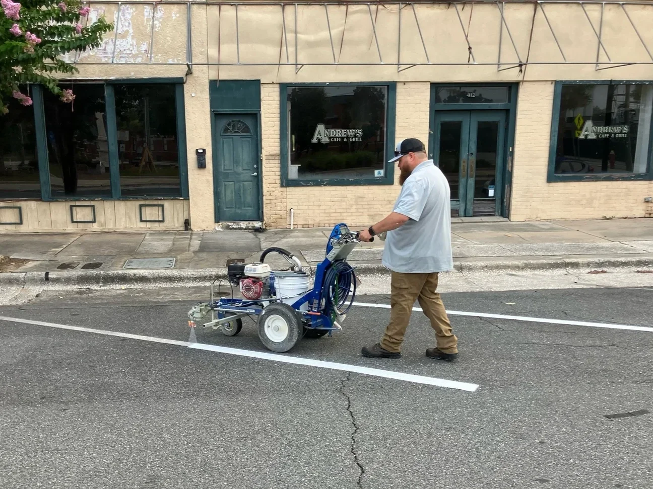 A man with a beard, wearing a white shirt, brown pants, and a baseball cap, is walking behind a small road marking machine on an urban street. There is a brick building with large windows and sign that reads 'A Andrews Cafe & Grill' in the background