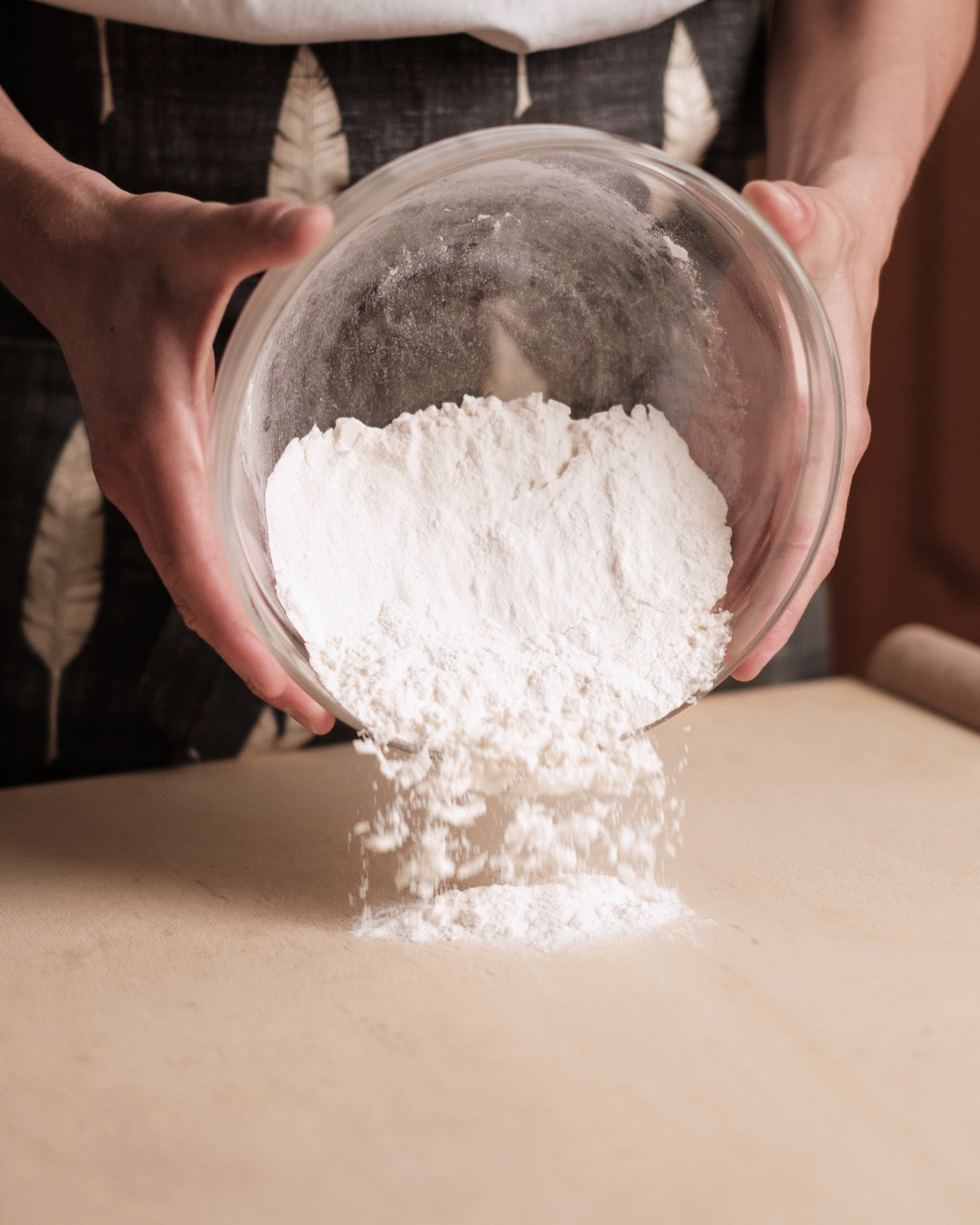 A person in an apron holds a  glass mixing bowl and tips flour onto a chopping board