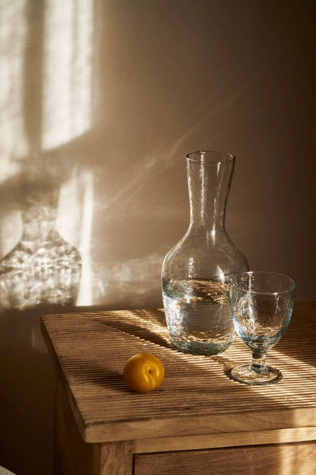 A clear glass carafe filled with water, accompanied by a matching glass, on a wooden surface with sunlight casting shadows and reflections.