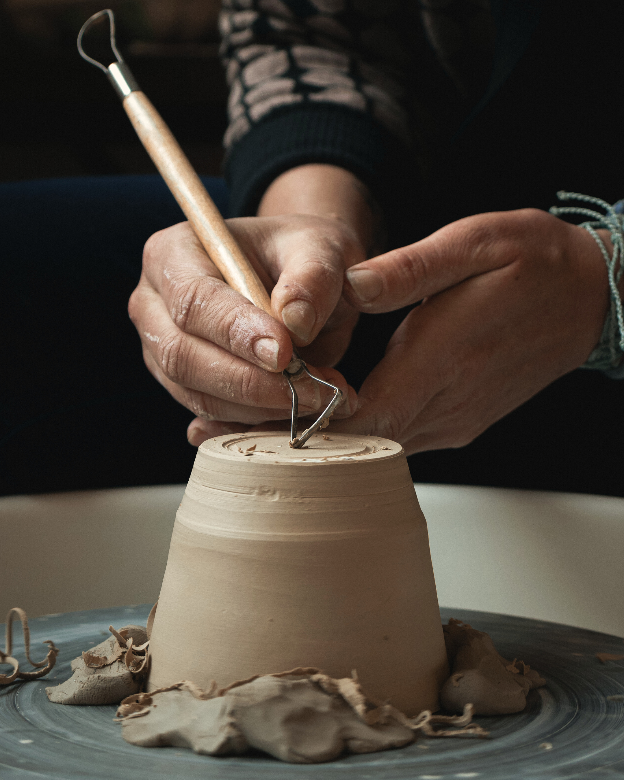 Hands shaping a clay pot on a pottery wheel with a tool.
