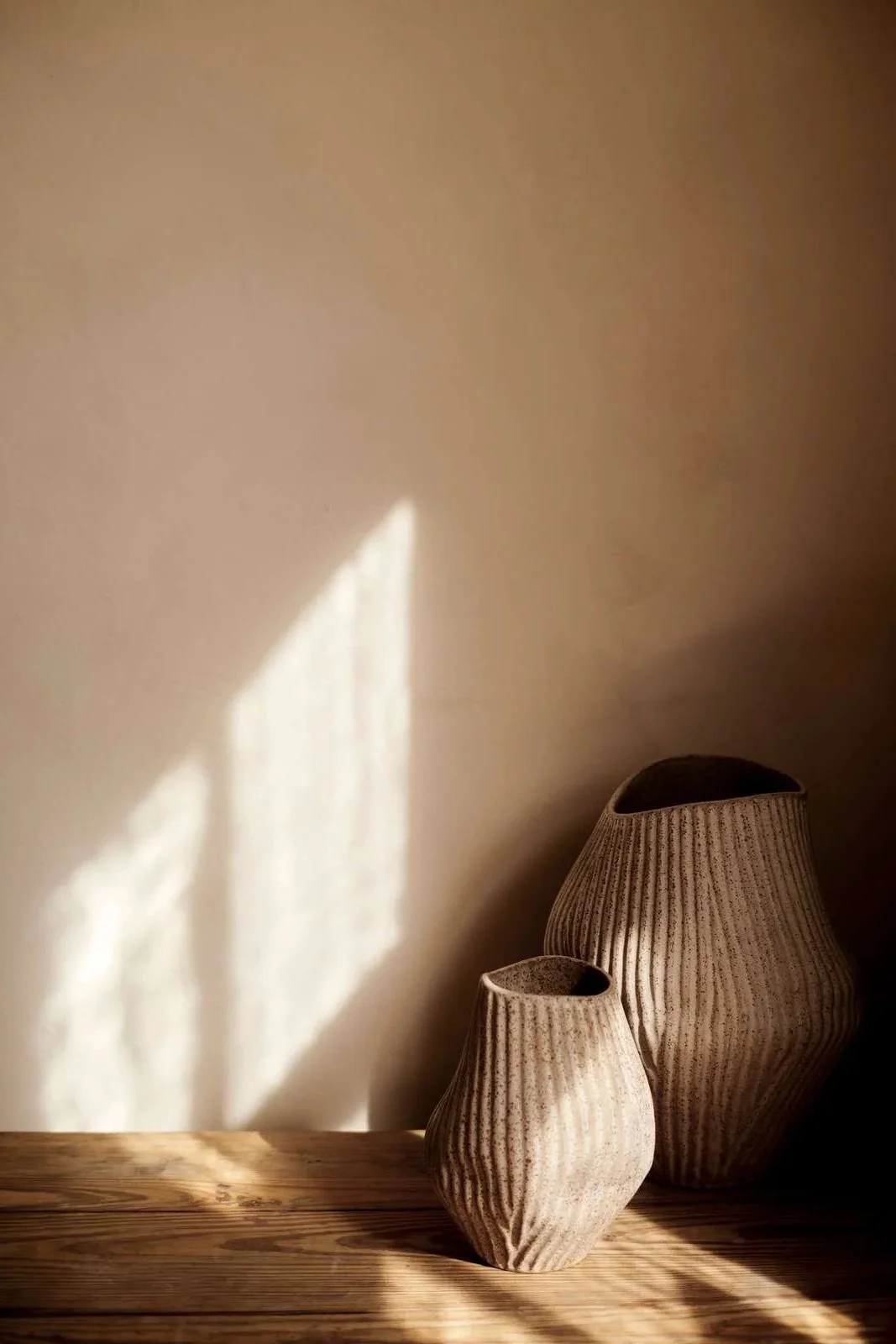 Two textured beige ceramic vases, one large and one small, sit on a wooden surface with sunlight creating a shadow on the beige wall behind them.