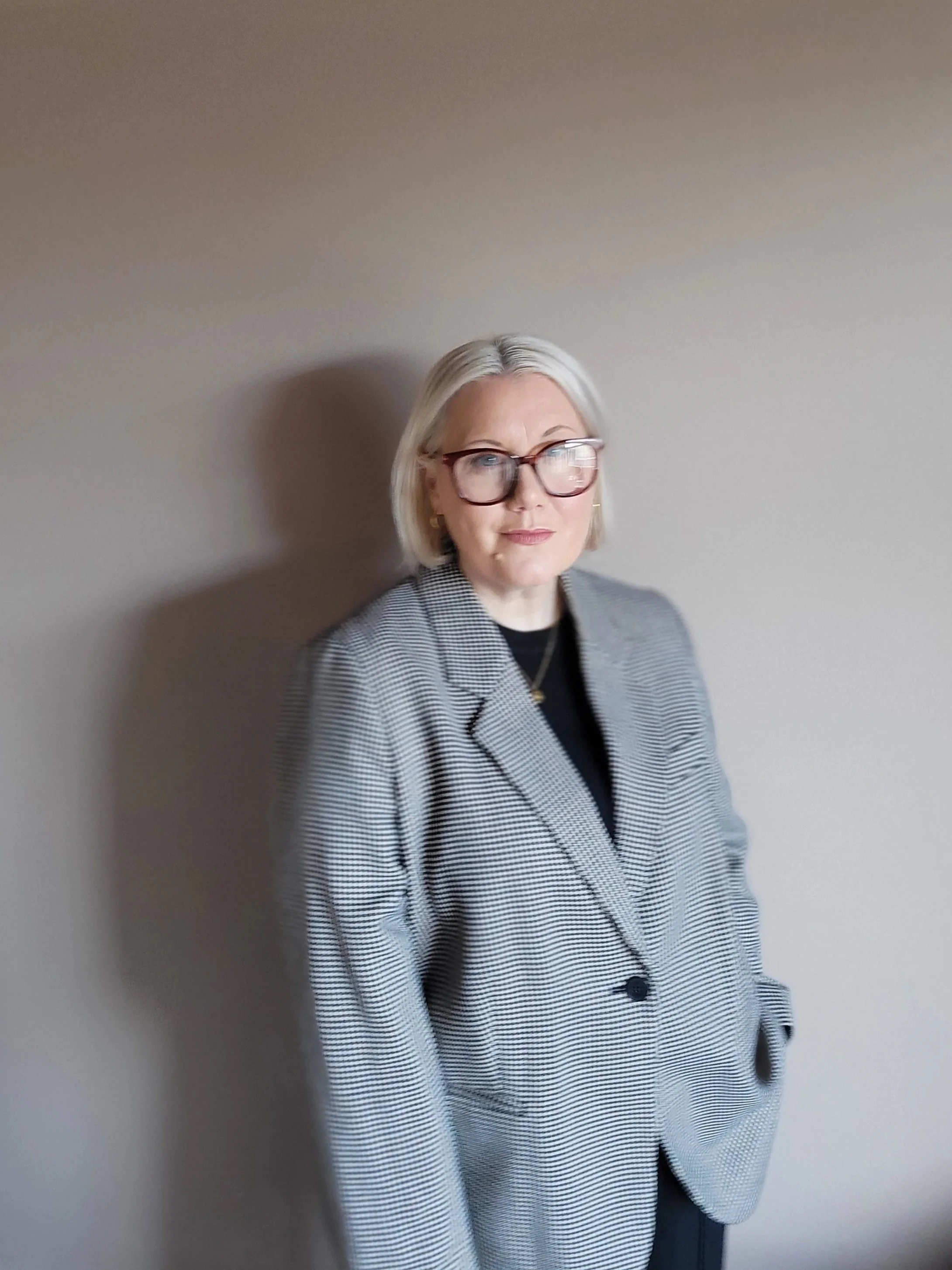A woman with blonde hair, wearing glasses, a gray checkered blazer, and a black top, standing against a plain beige wall.