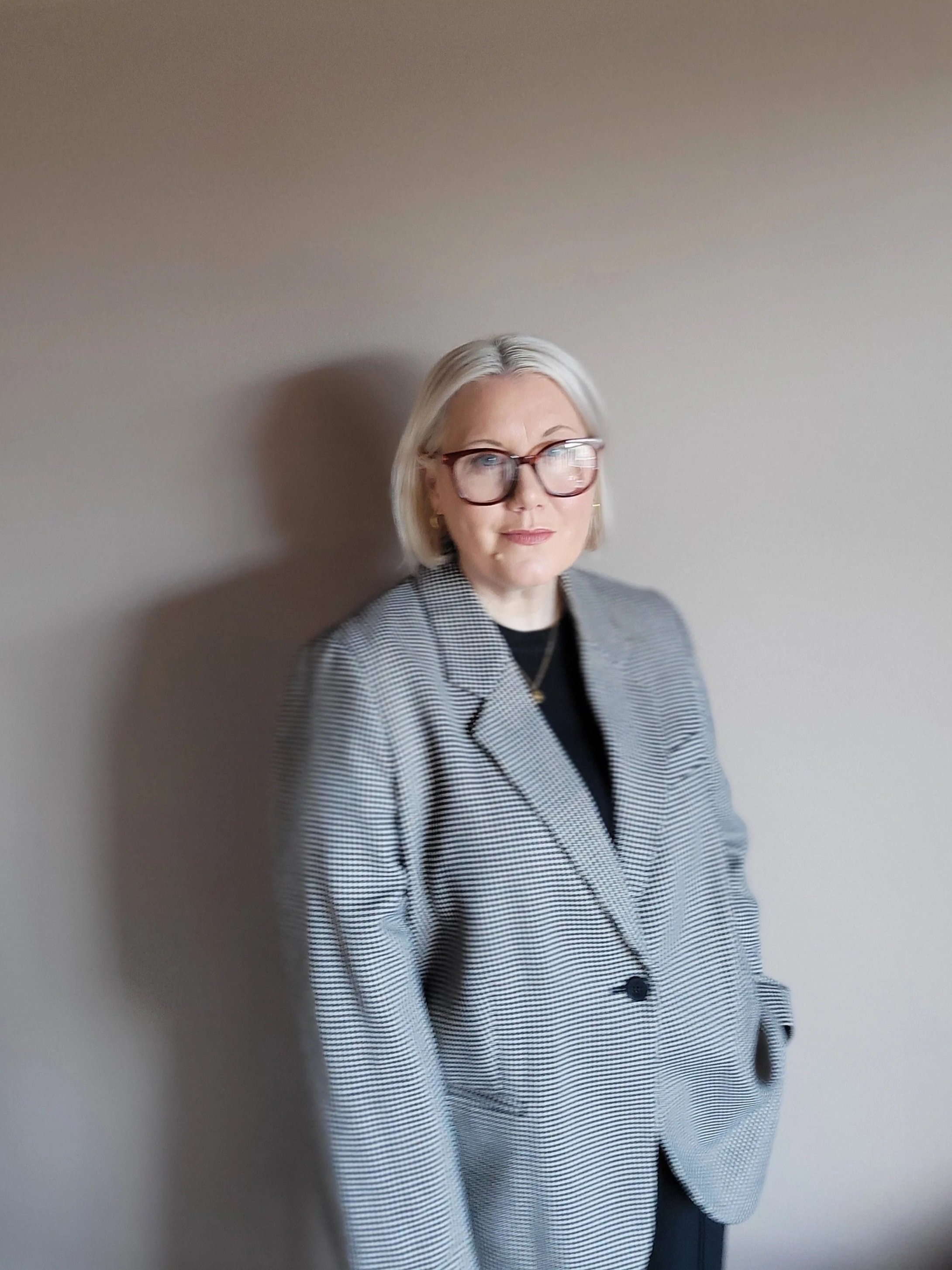 A woman with light hair and glasses wearing a gray checkered blazer and black shirt, standing against a plain wall.