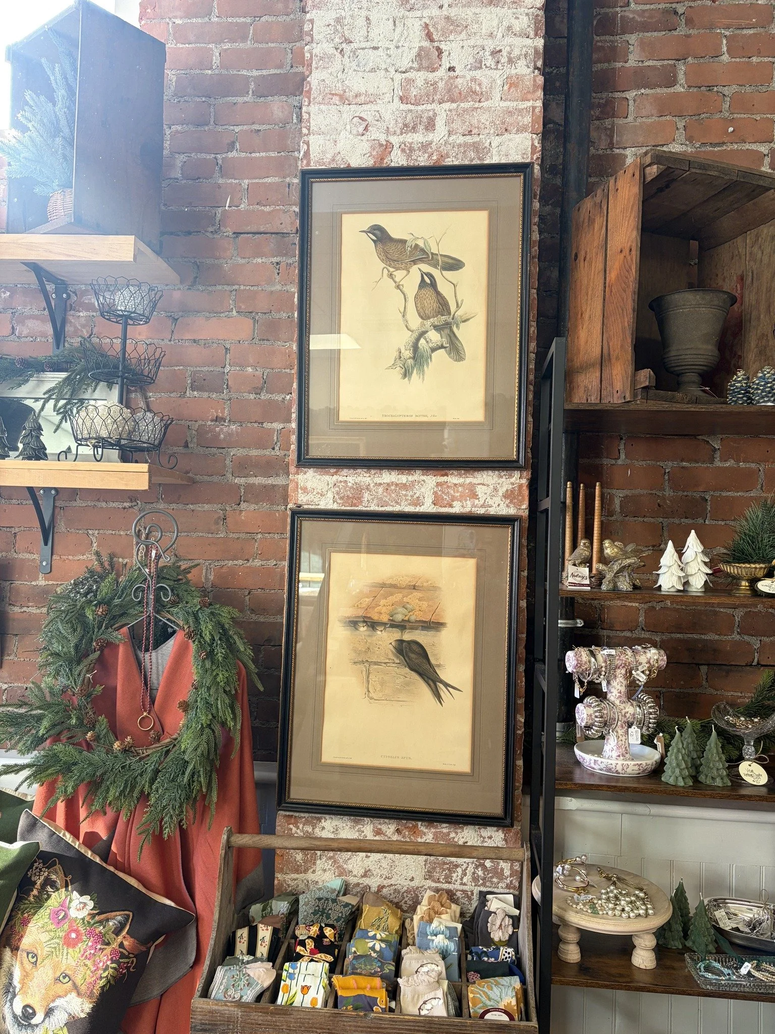 Decorative store interior with brick wall, framed bird illustrations, festive wreath, and various decorative items and jewelry displayed on shelves.