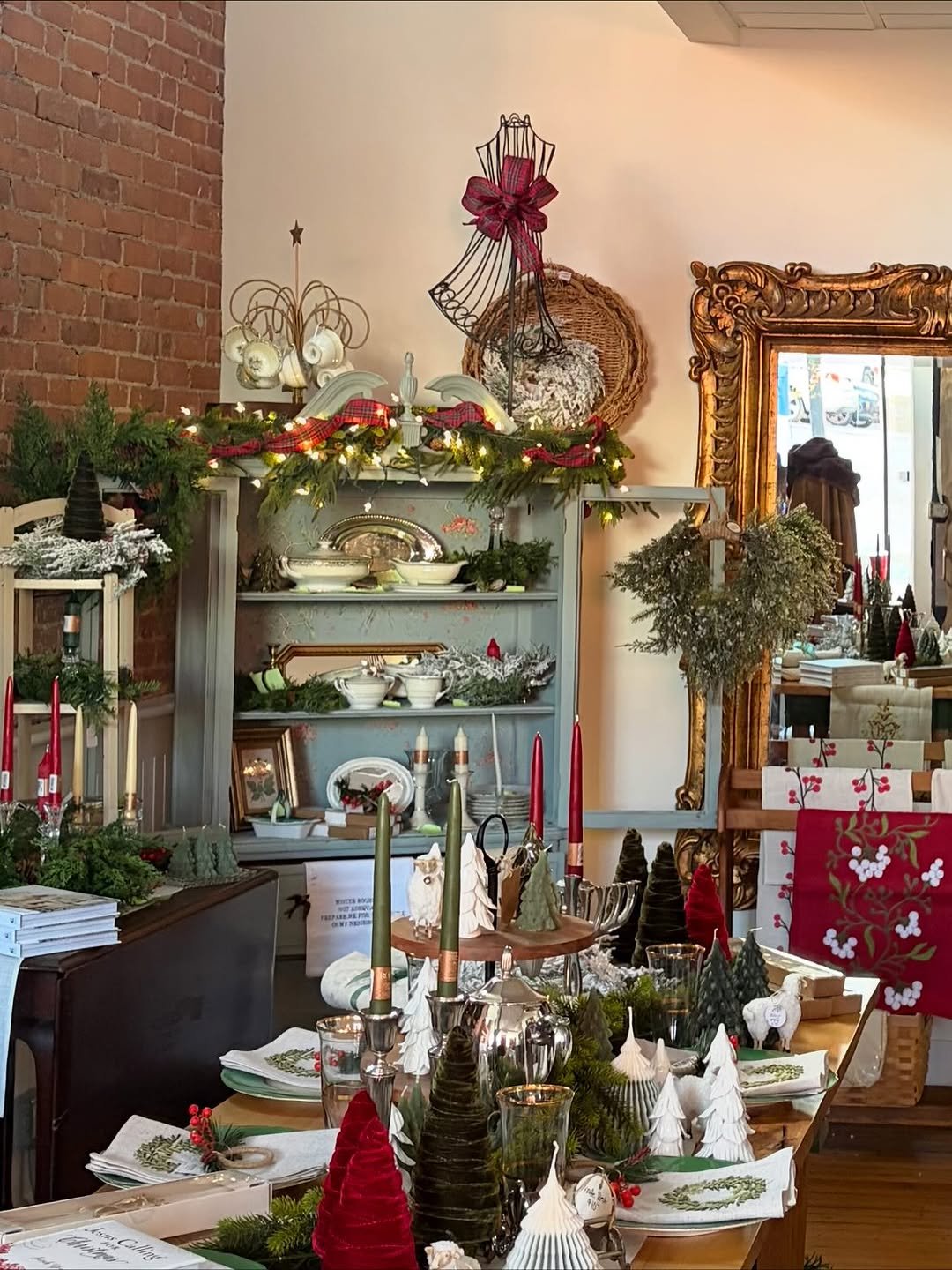 Festively decorated dining table and shelves with Christmas ornaments, including candles, trees, and greenery, inside a cozy room with a brick wall and ornate gold mirror.
