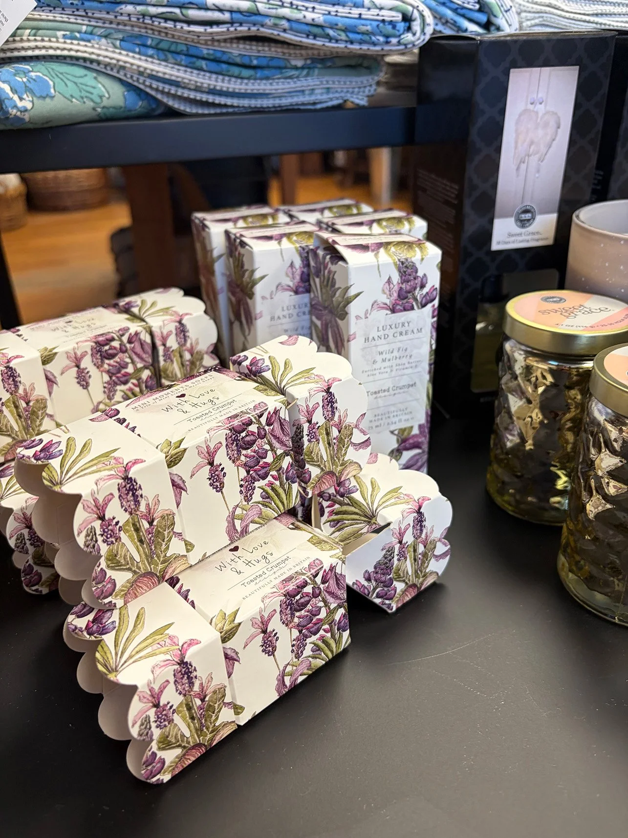 Festively decorated living room corner with a lit Christmas tree adorned with ornaments, a basket of holiday pillows, a wooden cradle with fabric rolls, dark green cabinet with holiday greenery and lights, and various wrapped gifts.