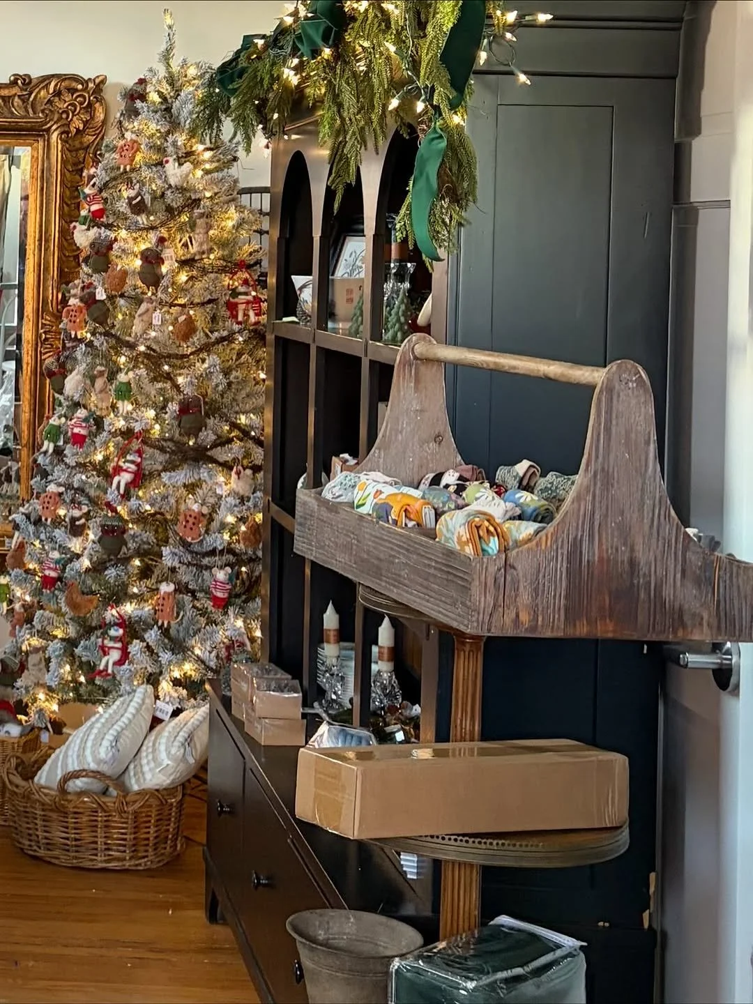 Festively decorated living room corner with a lit Christmas tree adorned with ornaments, a basket of holiday pillows, a wooden cradle with fabric rolls, dark green cabinet with holiday greenery and lights, and various wrapped gifts.