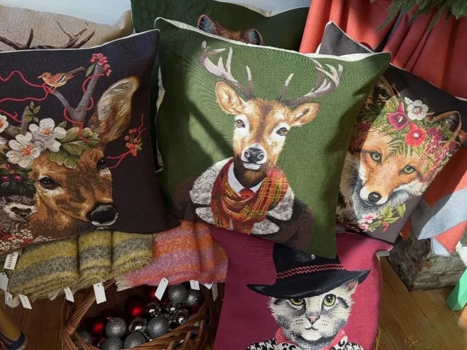 Decorative pillows with animal and holiday-themed embroidery, including a deer, a reindeer, a fox with flowers, and a cat with a witch hat, displayed on a surface with a basket of Christmas ornaments.