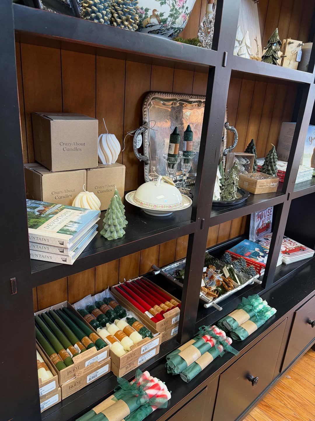 Decorative holiday-themed items including candles, ceramic trees, books, and wrapped candles on a black shelf with a wood-paneled back.