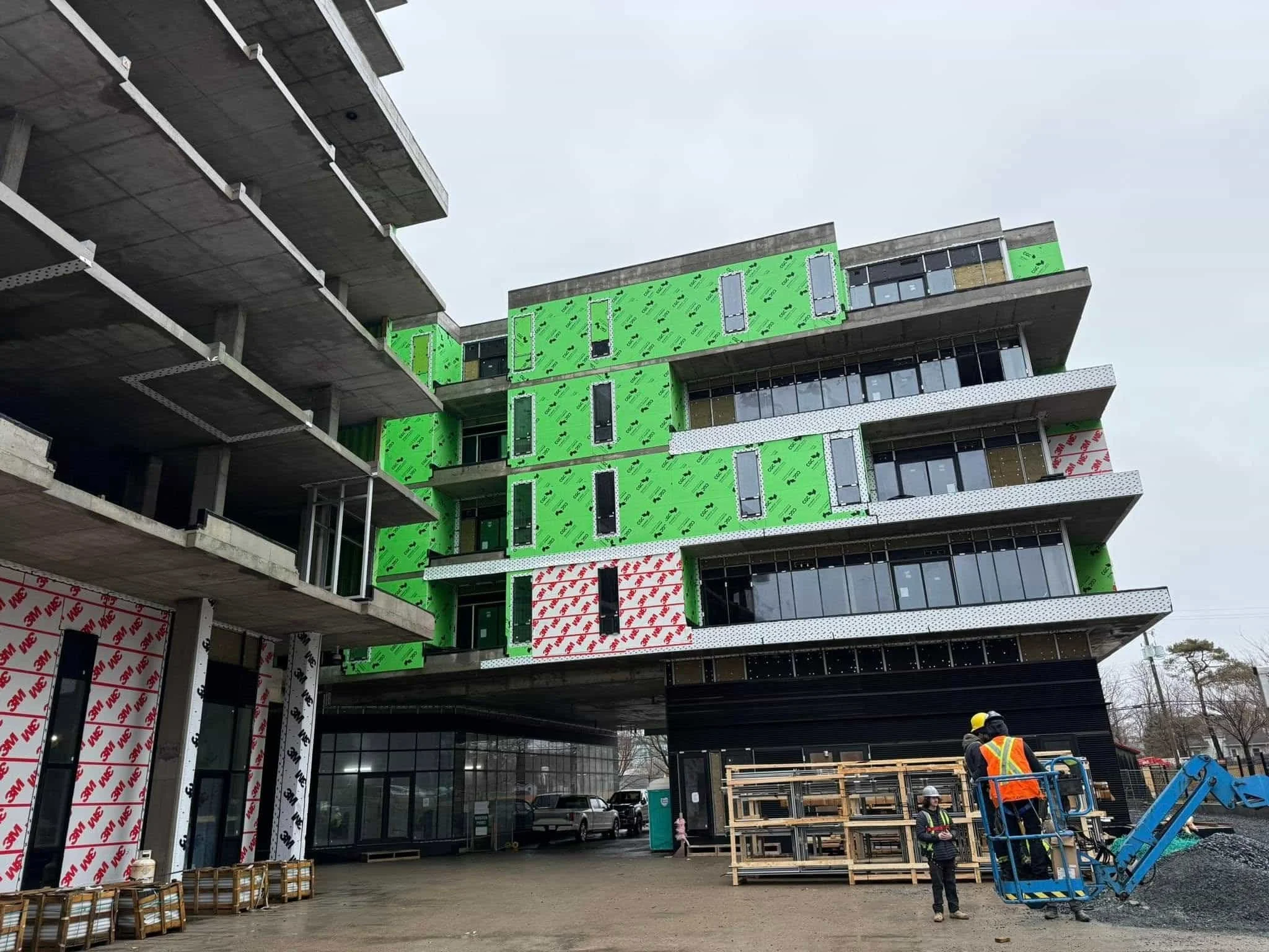 Construction workers in safety gear working on a multi-story building under construction with exposed concrete and green insulation panels.