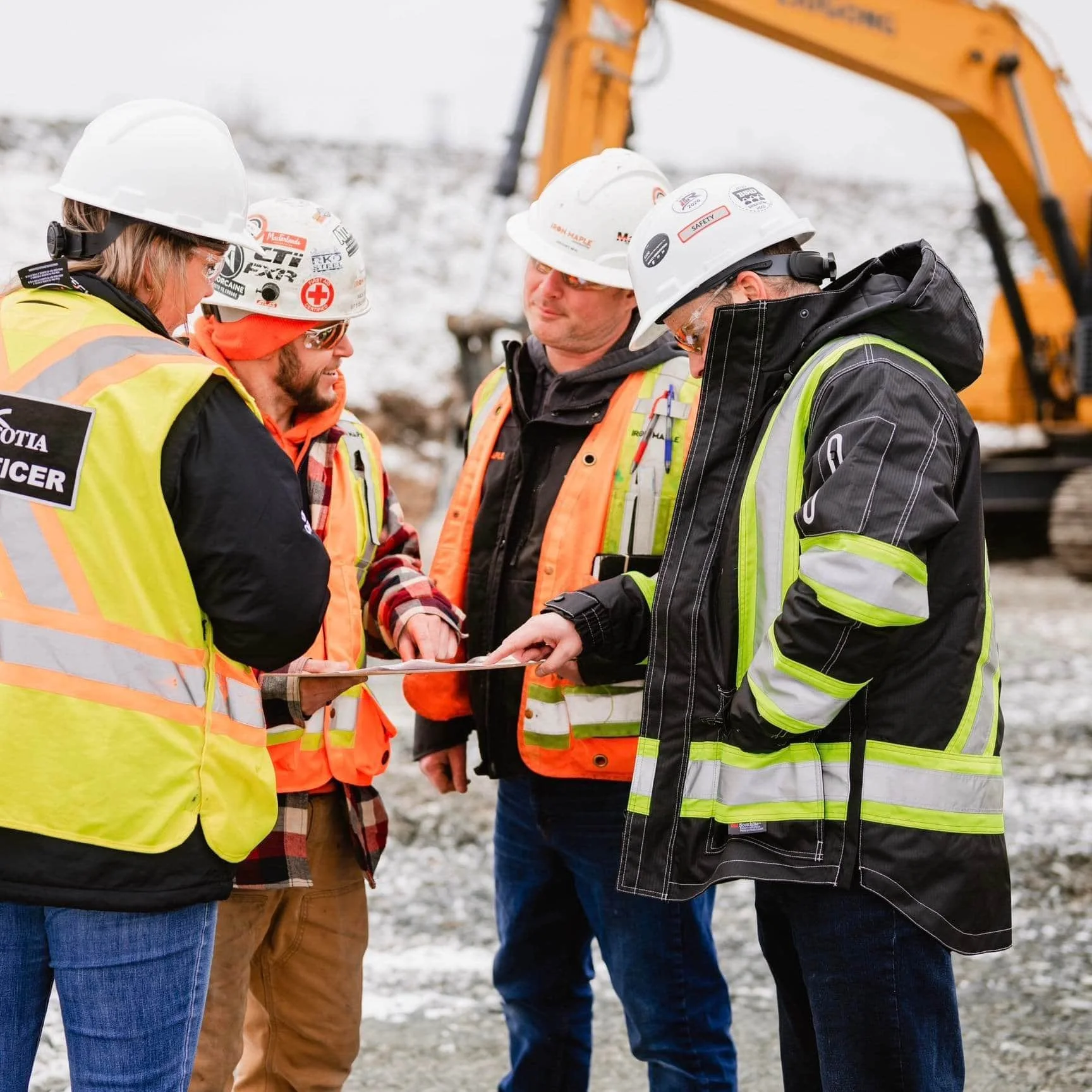 Group of construction workers and officials in safety vests and helmets reviewing plans at a construction site with equipment in the background.