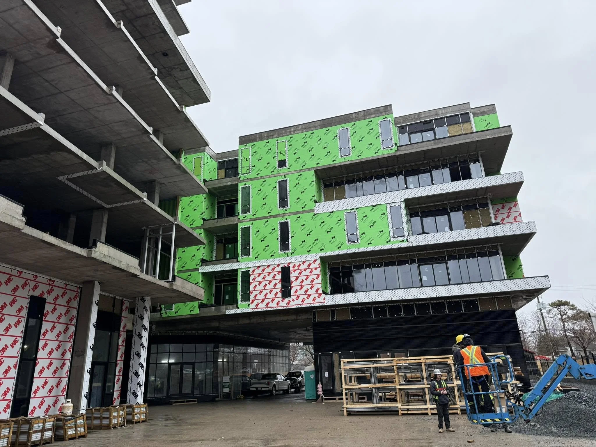 Under construction multi-story building with visible green insulation panels and black glass windows, construction workers in safety gear operating machinery and inspecting site, overcast sky.
