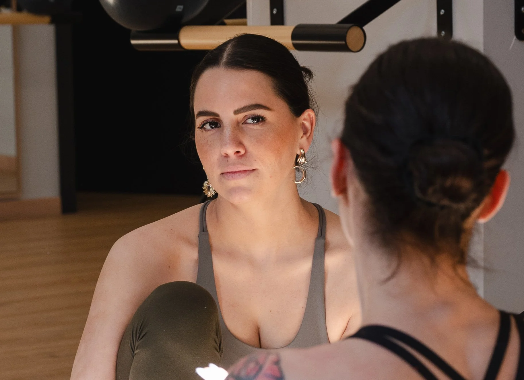 Dos mujeres conversando en un ambiente interior, una de ellas tiene cabello oscuro y usa aretes grandes, la otra tiene el cabello recogido en un moño y lleva tatuajes en el hombro.