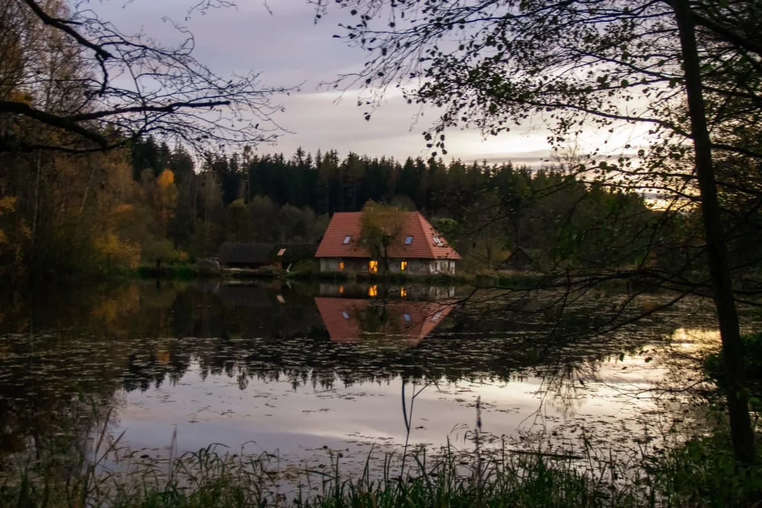 A peaceful house with a red roof and blue skylights sits by a pond in rural Czechia, with its reflection visible in the water. Trees with fall foliage surround the scene, and the sky is overcast during twilight.