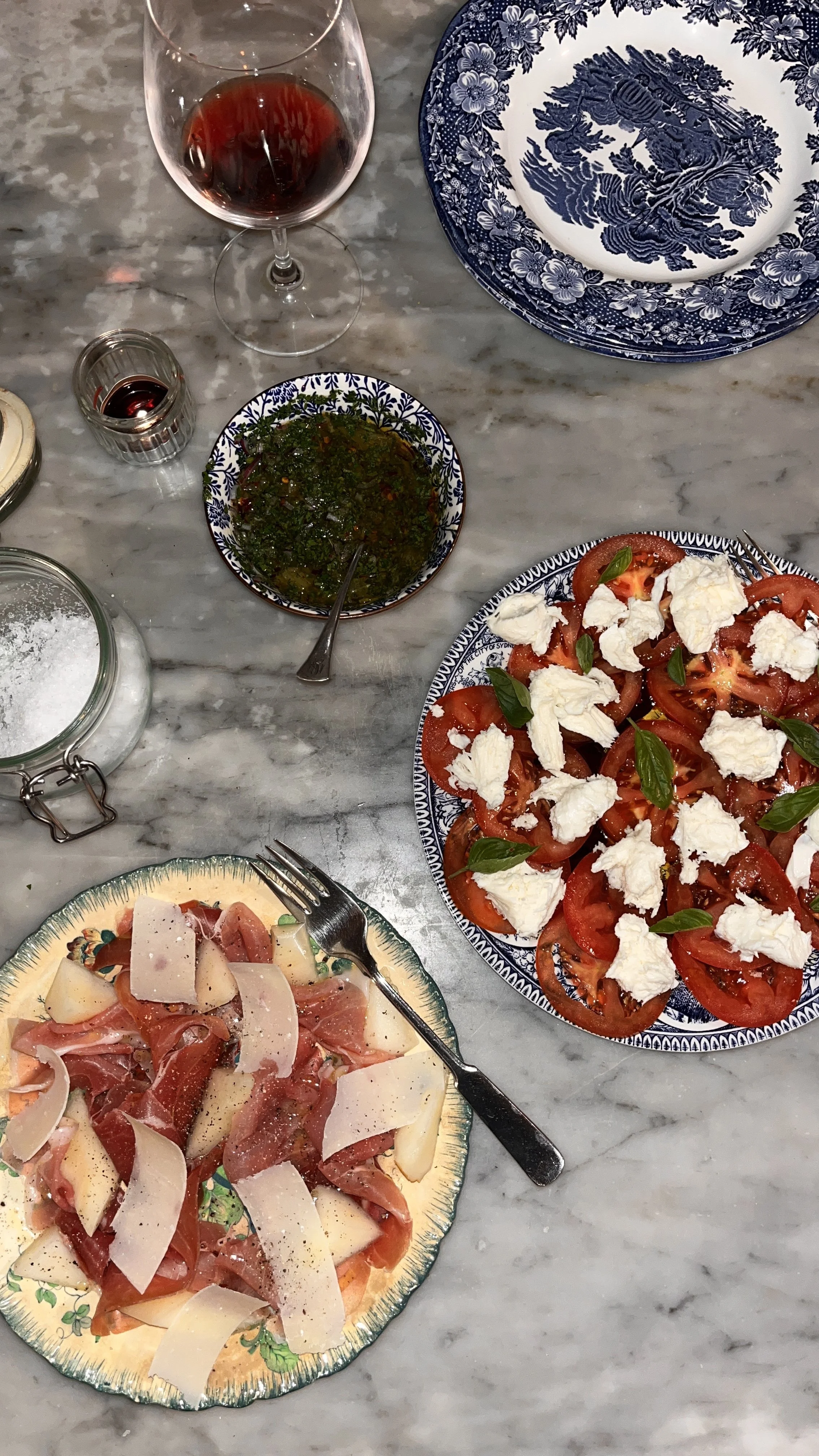 A marble table with a glass of red wine, a small shot glass with red liquid, a bowl of green sauce or dressing, a plate of sliced tomatoes with fresh basil and cheese, and another plate of sliced cured meats and cheese with a fork.