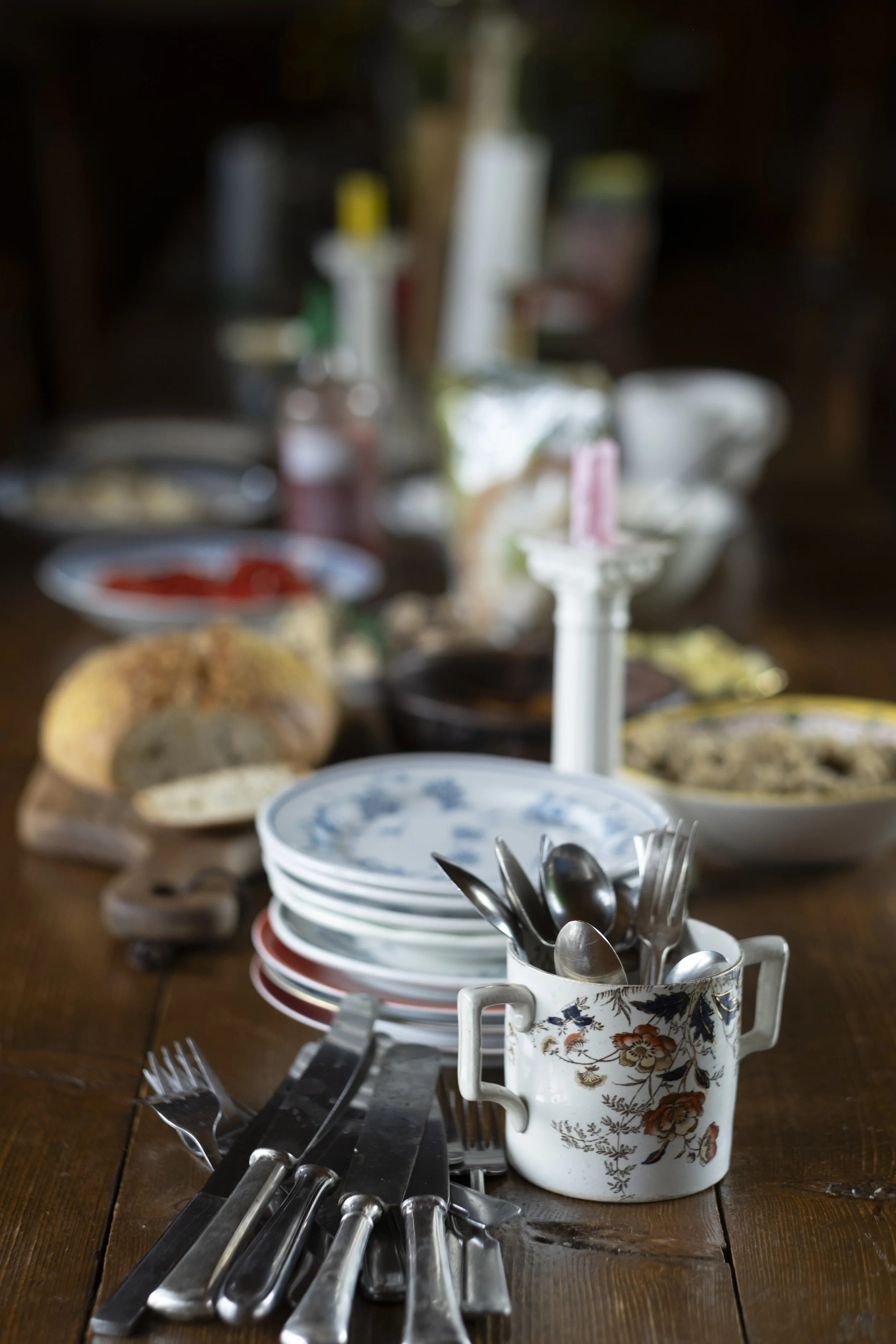 A rustic wooden table set for a meal with dishes, silverware, and condiments, with a stack of plates and a floral mug holding utensils in the foreground.