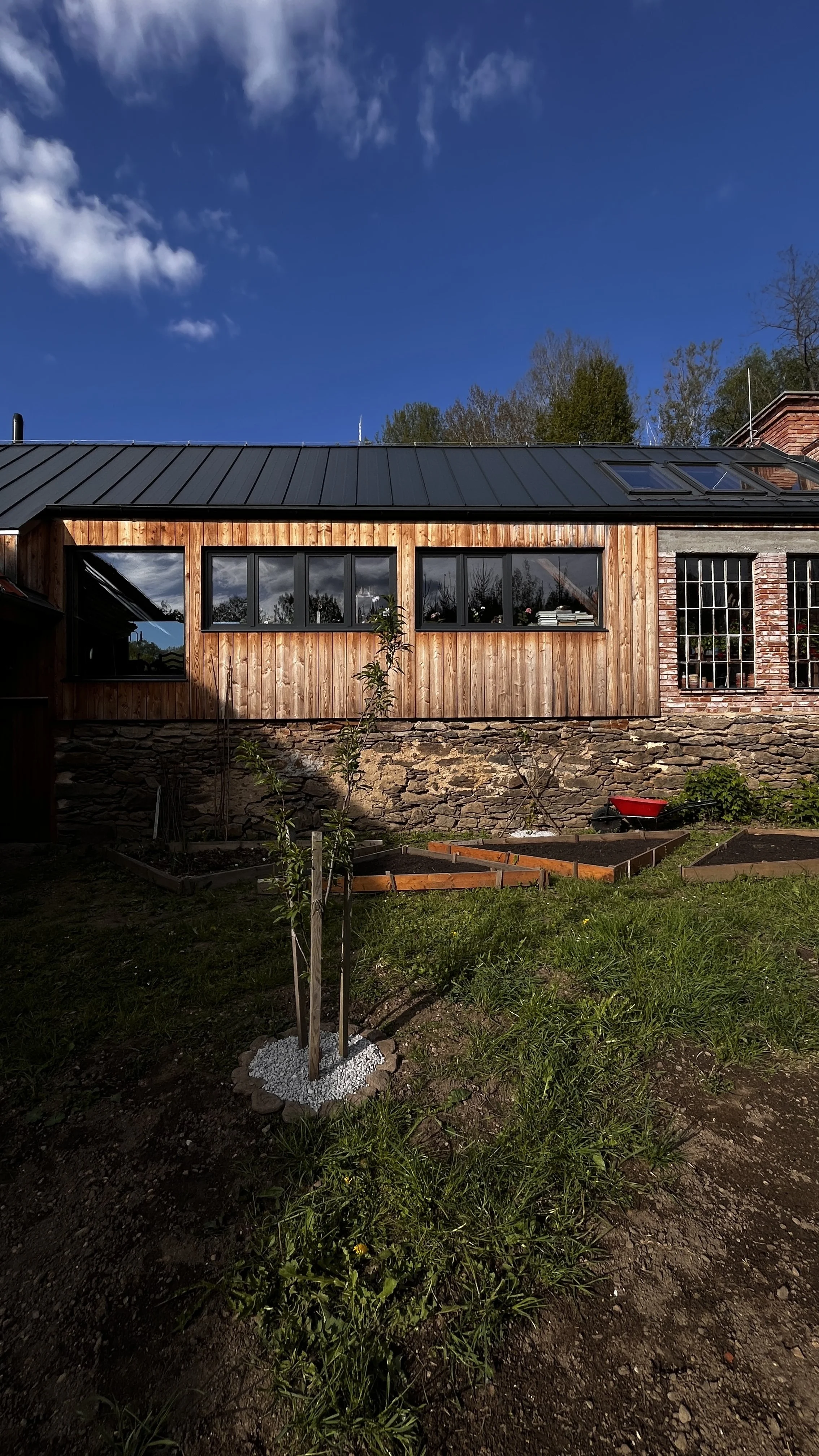 A house in the Czech countryside with a stone foundation, wooden siding, and large windows, under a blue sky with some clouds, with a garden in the foreground.