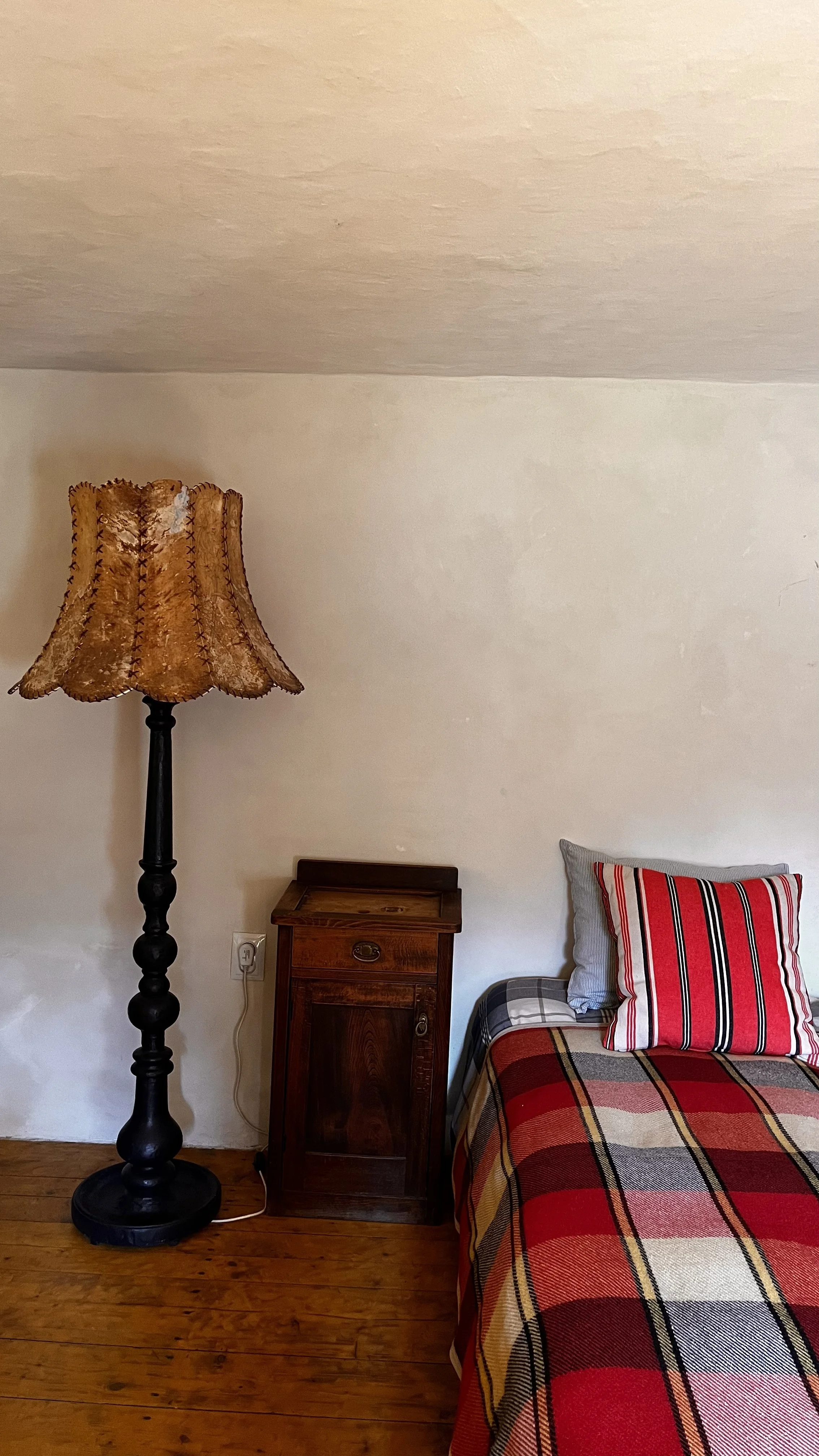 A bedroom corner with a tall wooden floor lamp with a fringed fabric lampshade, a small wooden nightstand, and part of a bed with a red, yellow, gray, and black plaid blanket and a striped pillow.