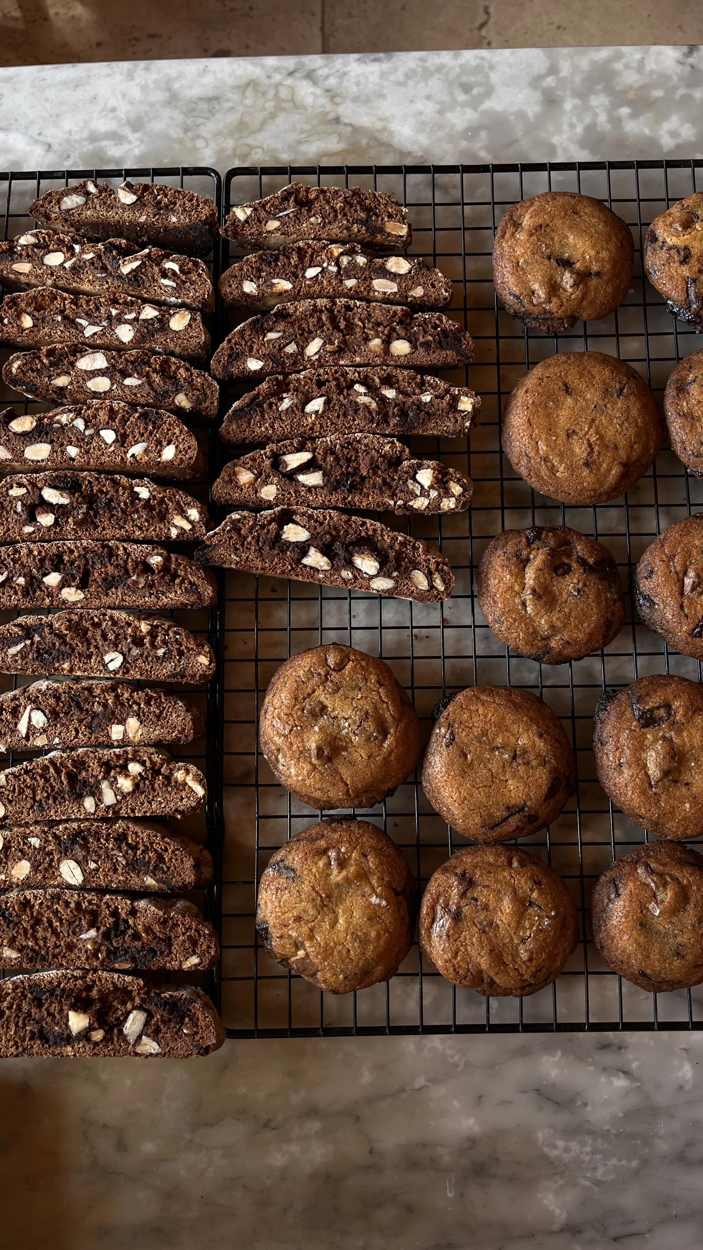 Chocolate biscotti with almonds on a cooling rack next to chocolate chip muffins on a marble countertop.