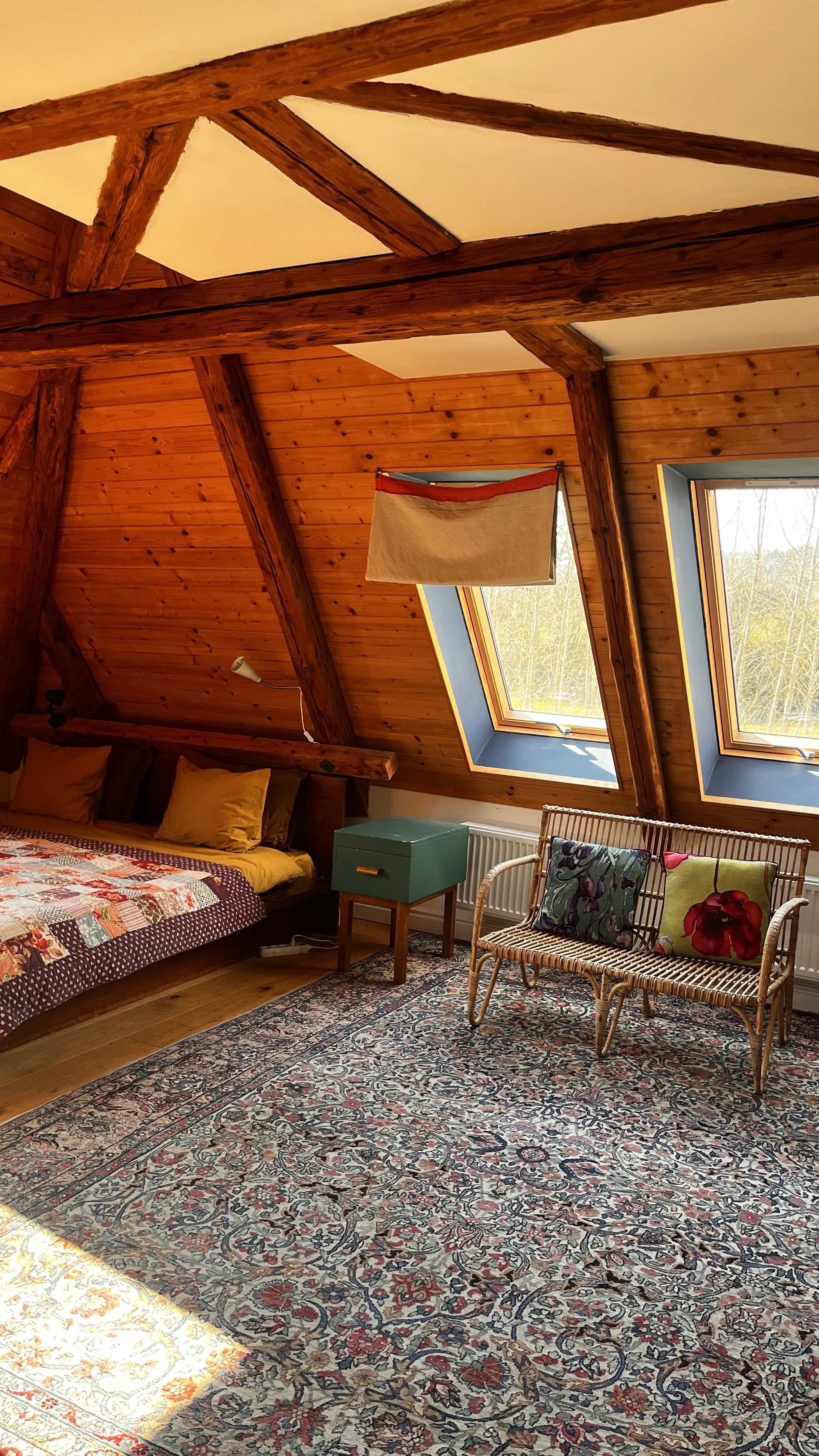 A cozy attic bedroom with wooden walls and ceiling beams, two skylight windows, a bed with yellow pillows and a patchwork quilt, a small green nightstand, a rattan sofa with decorative pillows, and a patterned area rug.