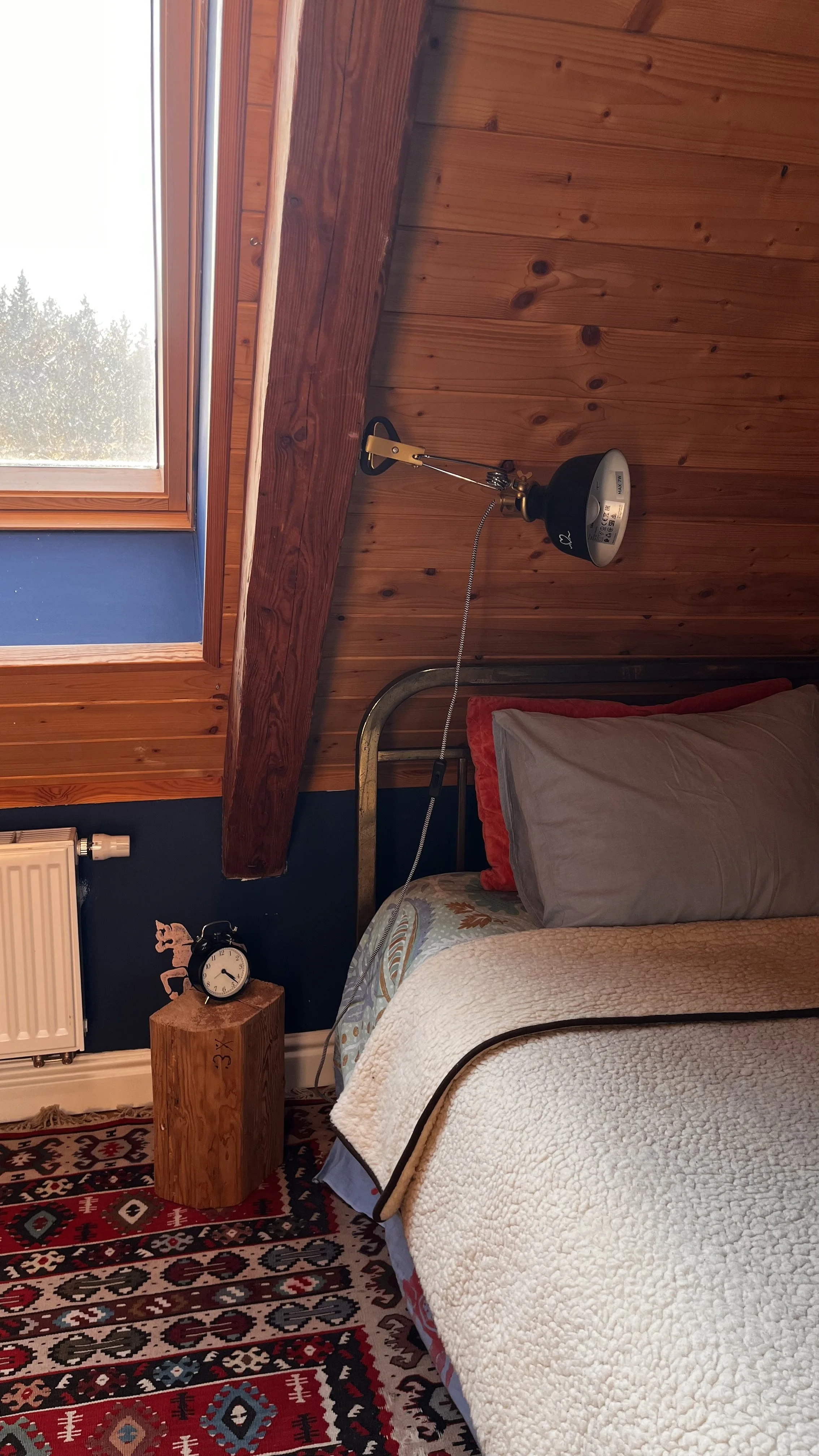 A cozy bedroom with a wooden ceiling and wall paneling, featuring a window with a view of trees, a bed with gray and red pillows, a wooden nightstand with a clock and decorative horse, and a wall-mounted lamp.