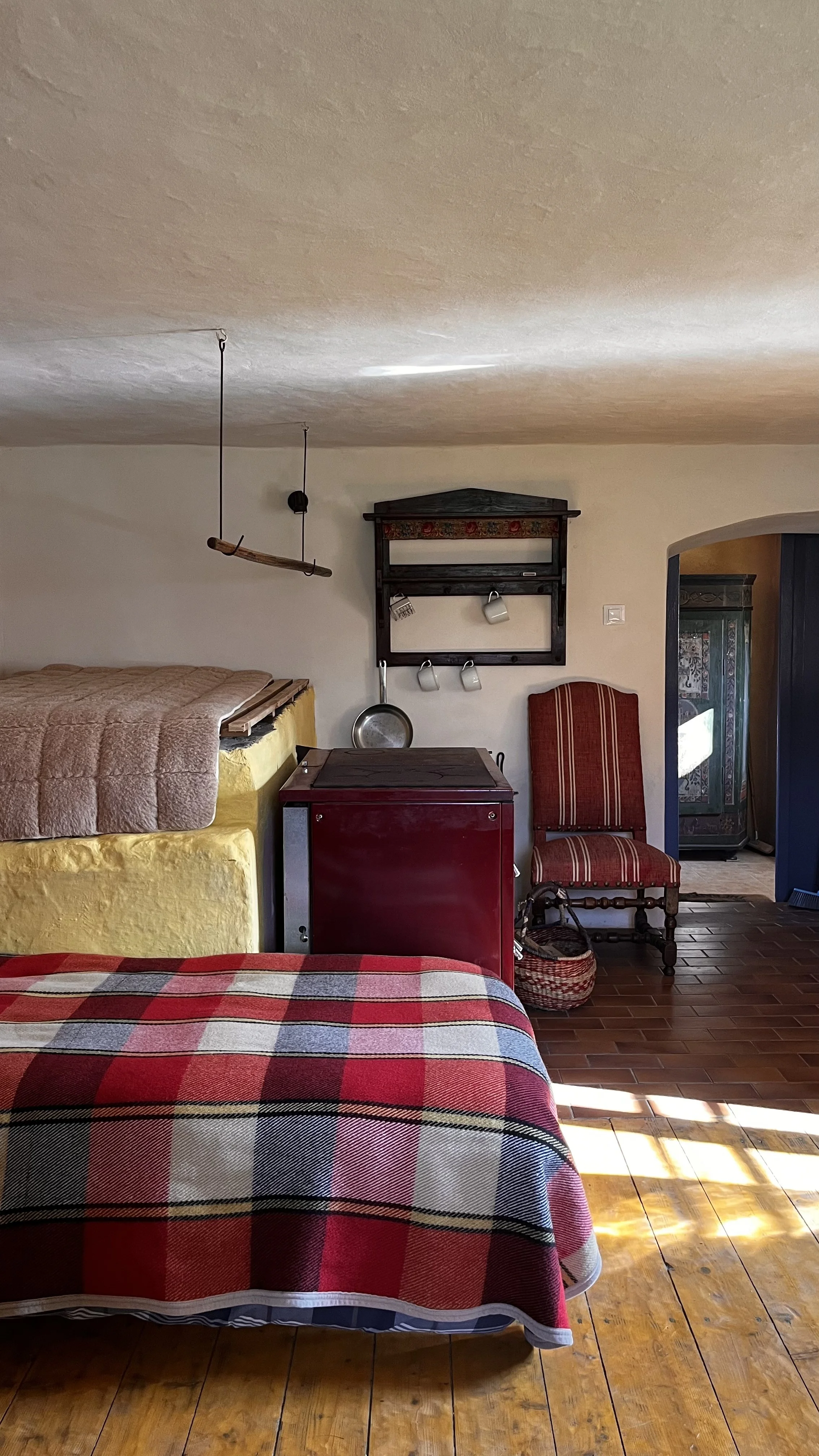 Interior of a rustic bedroom with a bed covered in a red and gray plaid blanket, a red cabinet, and a vintage chair with red striped upholstery. There are hanging cups, an old wooden shelf, and a swing hanging from the ceiling. The room has wooden floors and a doorway leading to another room.