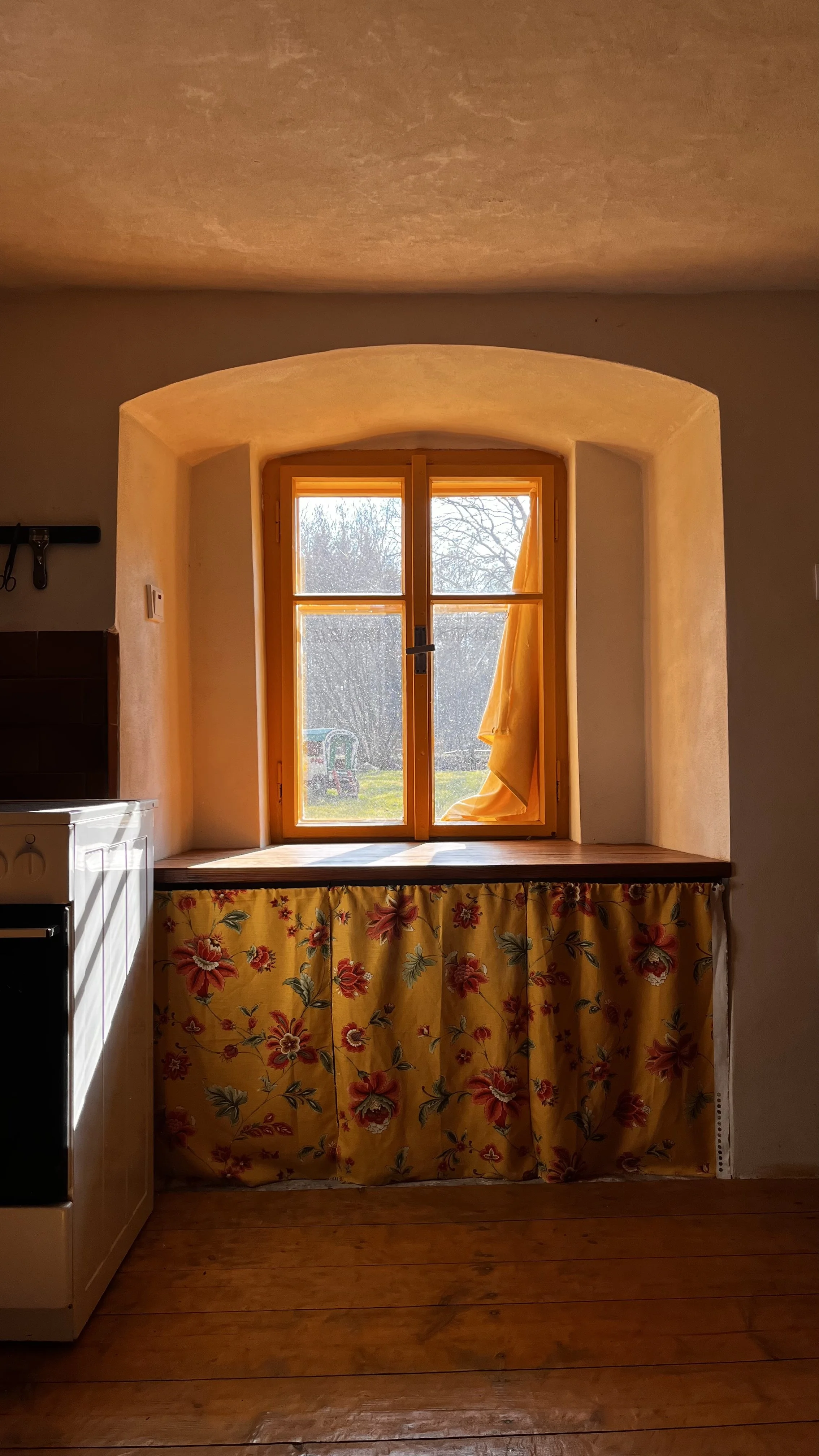 A bright window with a yellow curtain partway pulled aside, revealing an outdoor scene with trees and a small green outdoor play structure. Under the window, there is a floral curtain with red, pink, and gray flowers on a yellow background, hanging down over a small space.
