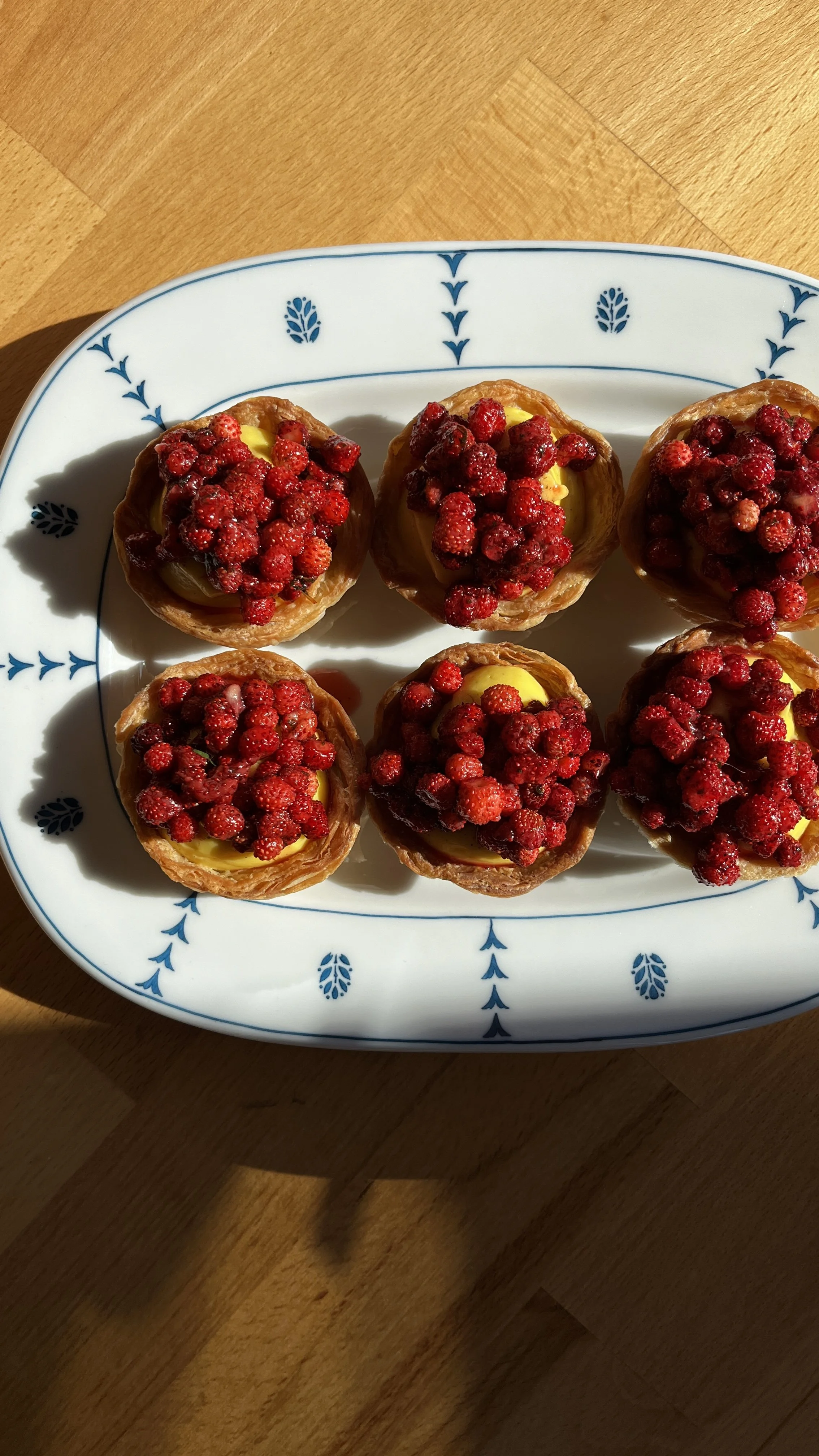Eight small pastries topped with red berries arranged on a white plate with blue designs, resting on a wooden surface.
