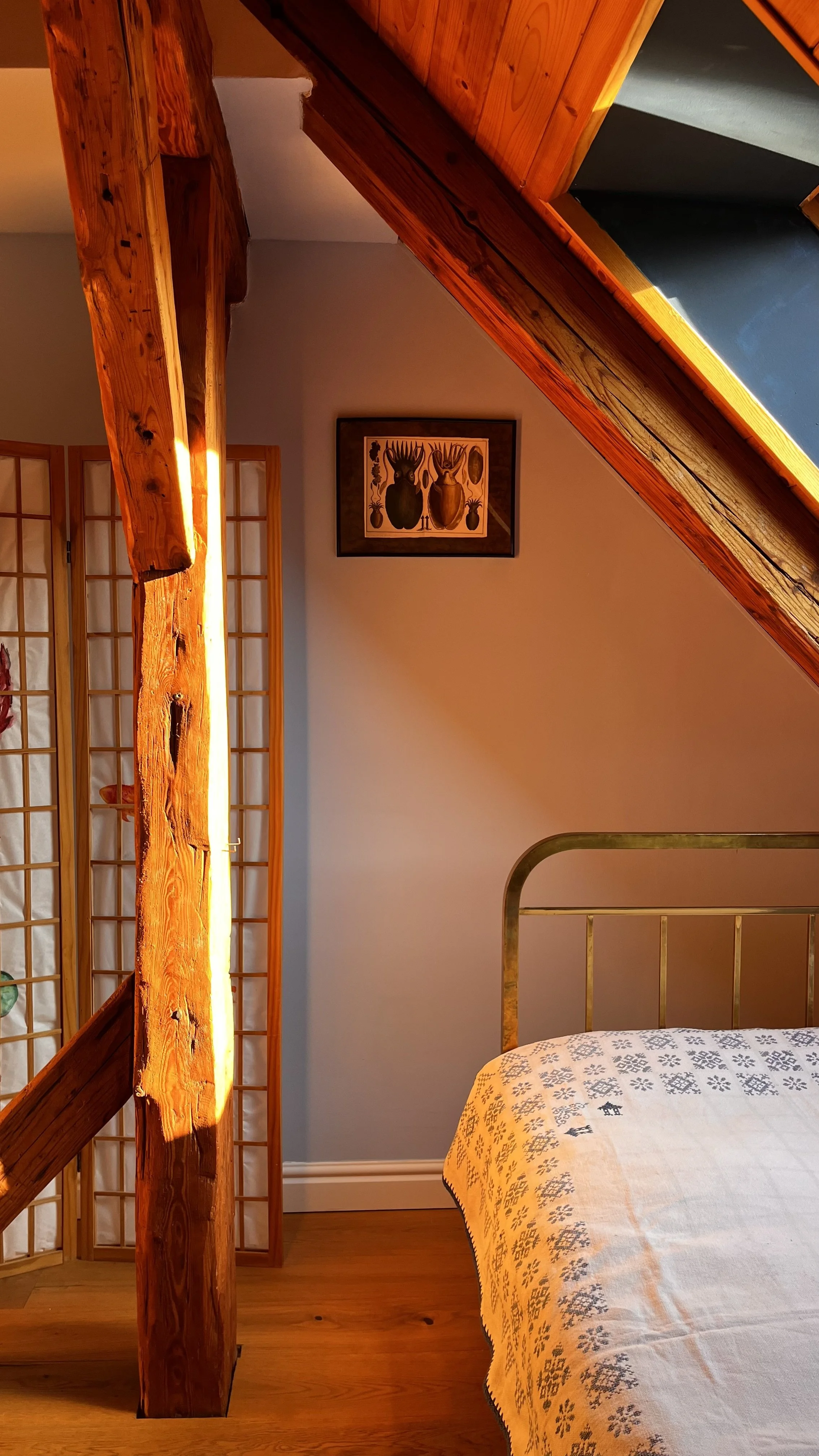 A cozy bedroom corner with a slanted ceiling, wooden beams, a metal bed frame, and a white bedspread with blue floral patterns. A wooden room divider and framed artwork of abstract animal figures hang on the wall.