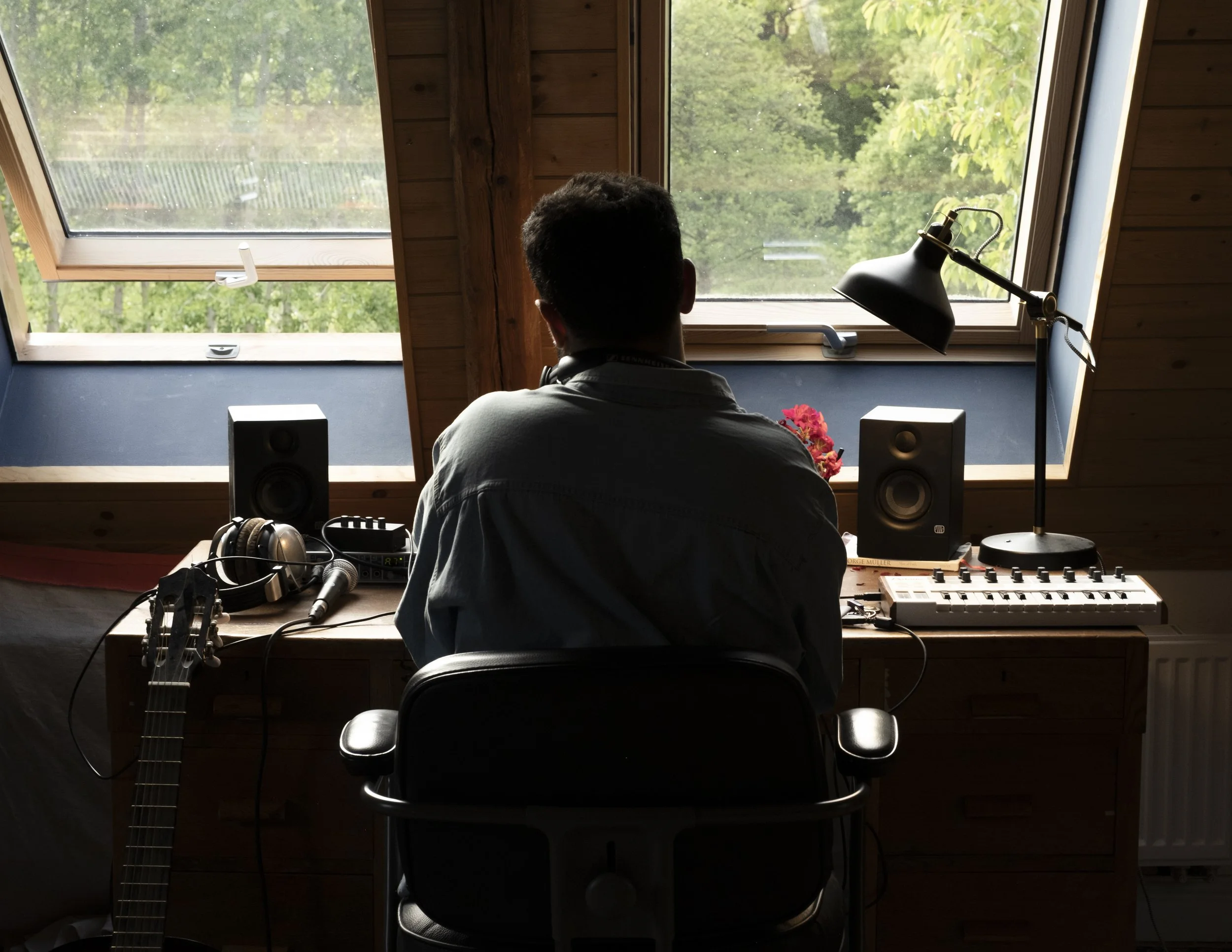 Person sitting in a home music studio, facing a window with a view of trees, with audio equipment and a guitar on the desk.