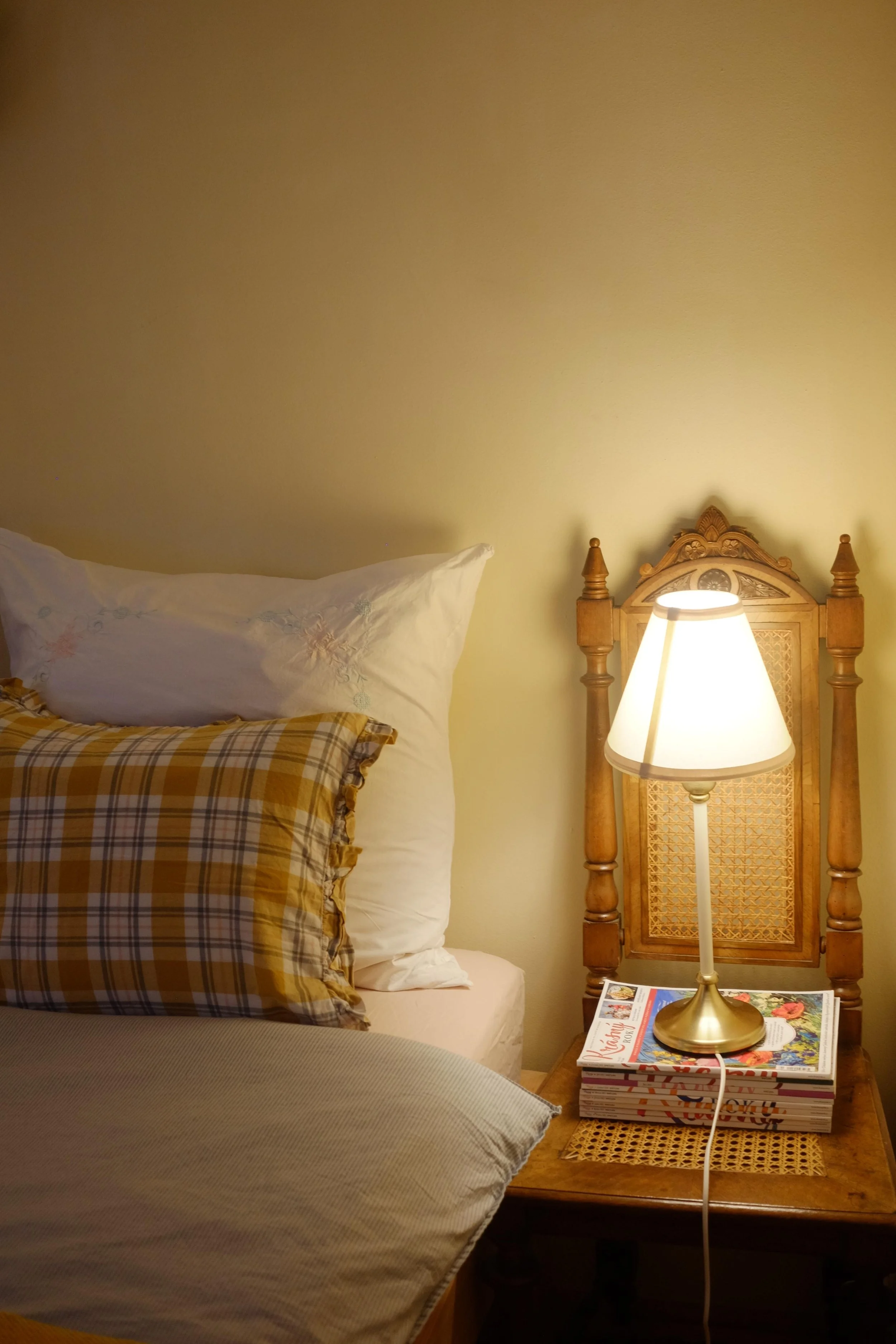 A cozy bedroom scene with a bed, a wooden nightstand, a bowl lamp, and a stack of magazines. The headboard is wood with decorative carvings, and the bed has white and plaid pillows.