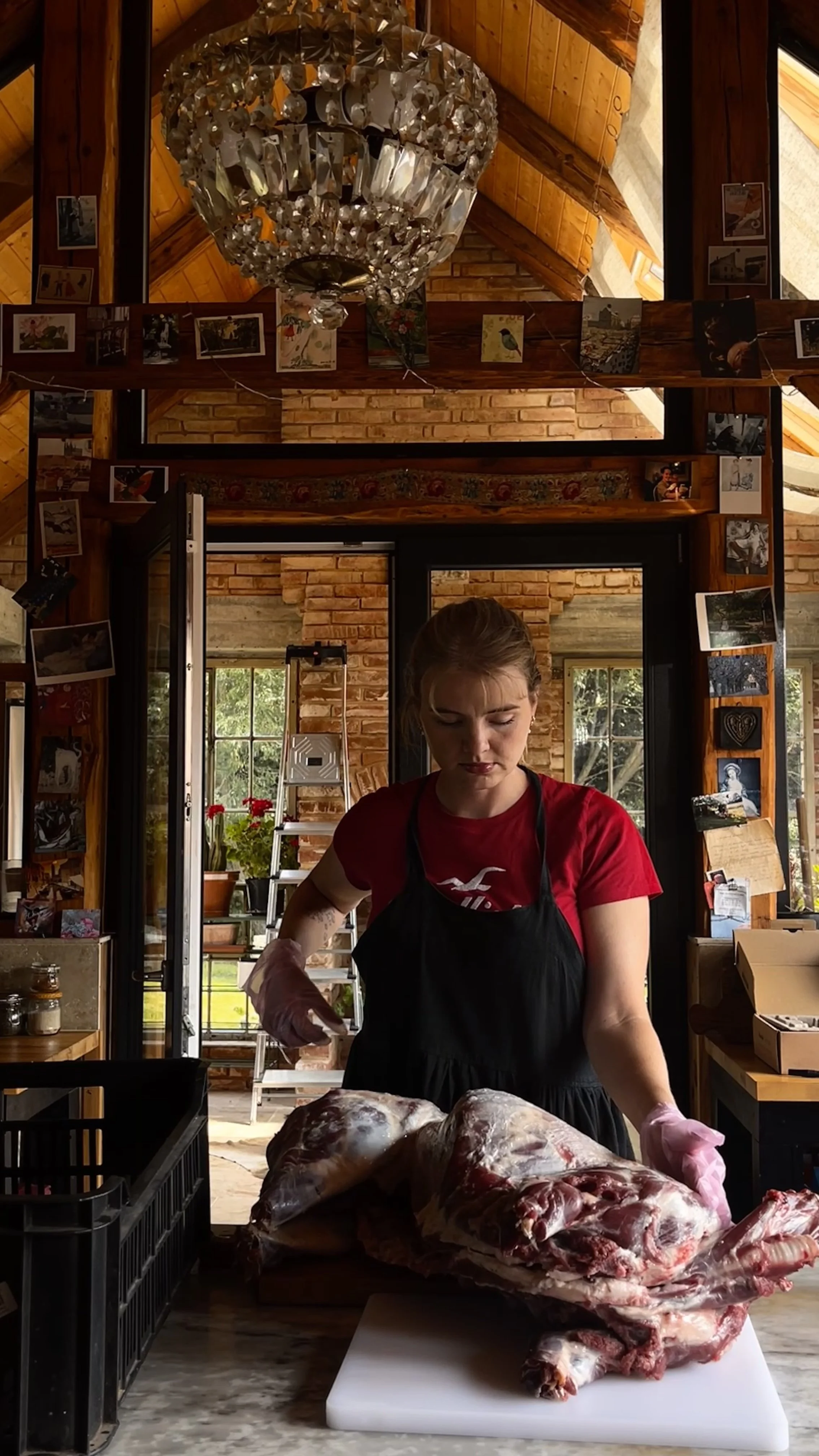 Young woman in black apron and pink gloves butchering a large animal carcass in a rustic kitchen with a chandelier overhead and photos hanging on the wooden beams.