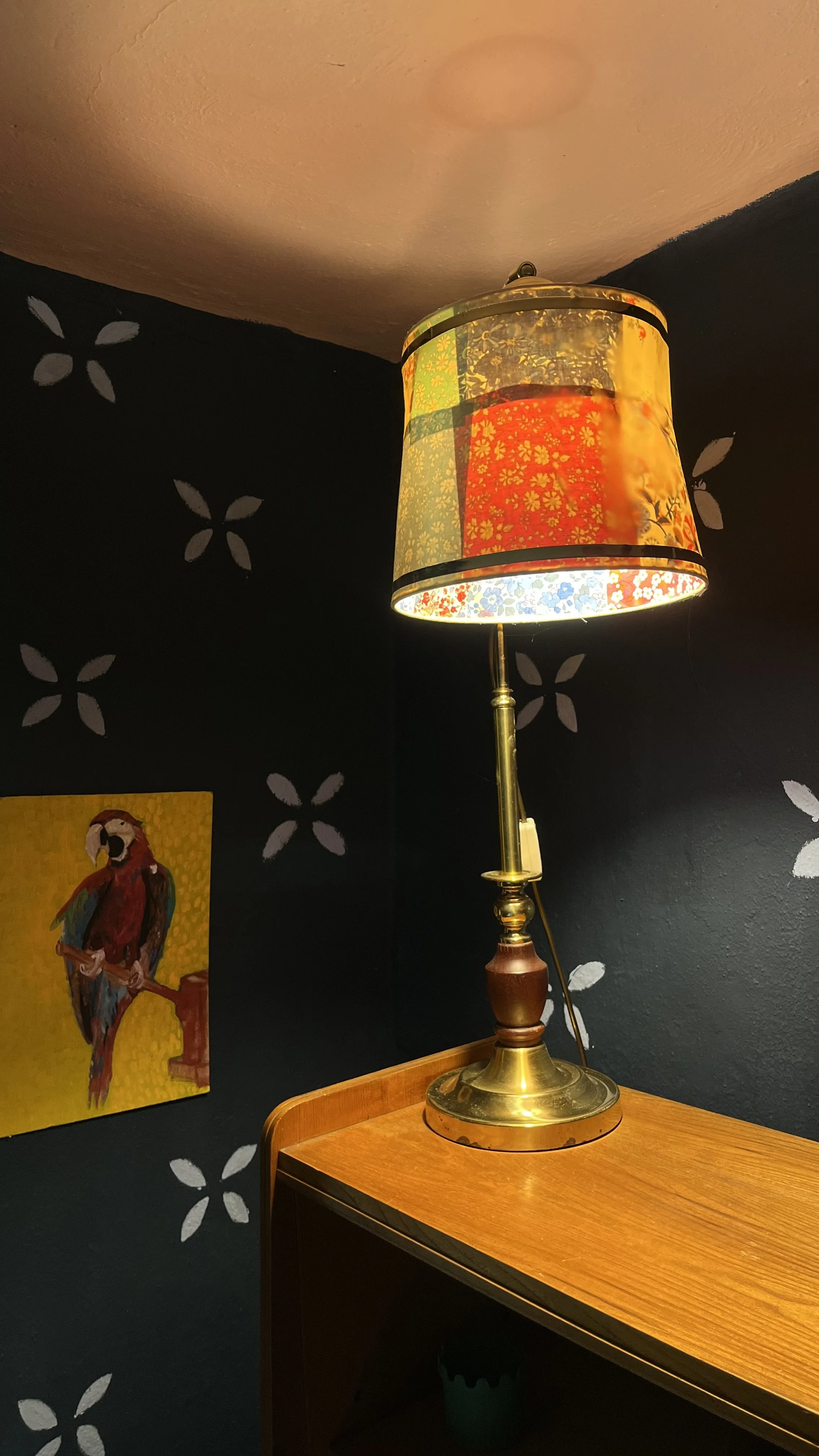 A table lamp with a colorful patchwork fabric lampshade, standing on a wooden table in a room with dark walls decorated with white flower patterns and a small painting of a parrot.
