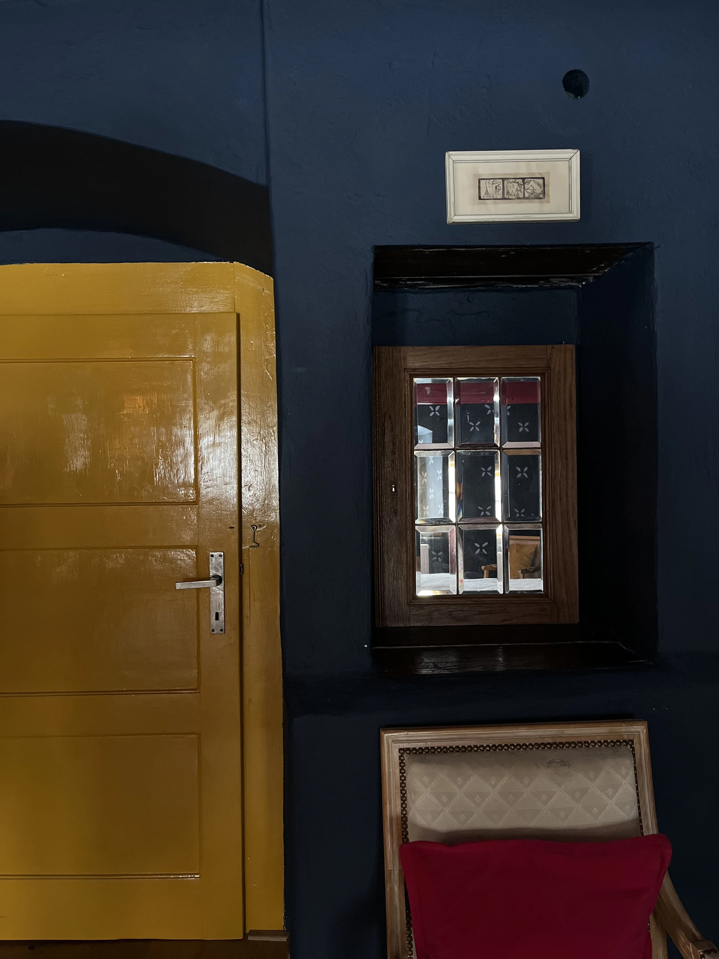 Interior of a room with a yellow door, dark blue walls, a small window with stained glass, a framed picture above the window, and a beige chair with a red cushion.